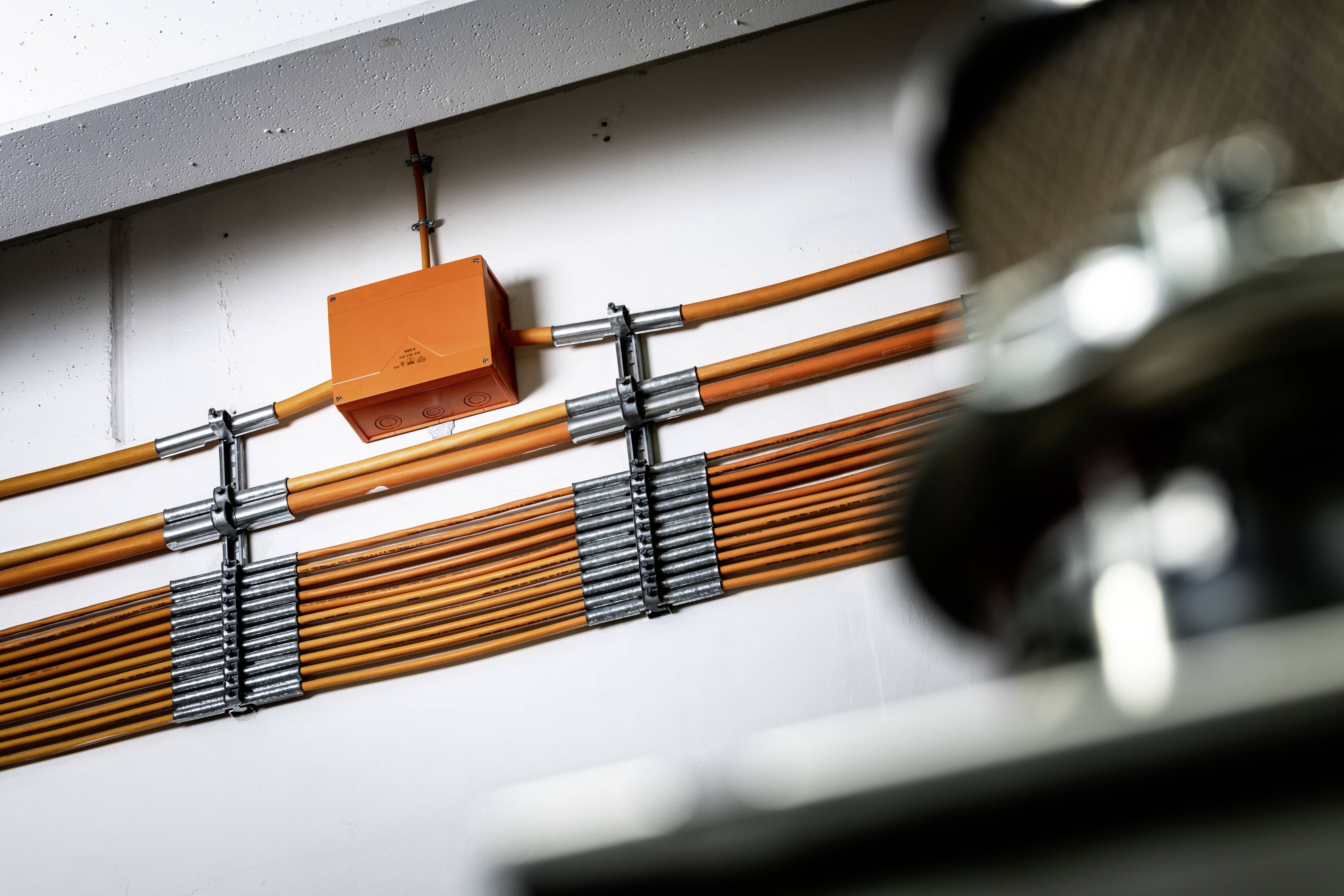 Ein oranges Kabelsystem an einer Wand, organisiert mit Halterungen. Im Vordergrund ist ein unscharfes Metallobjekt erkennbar.