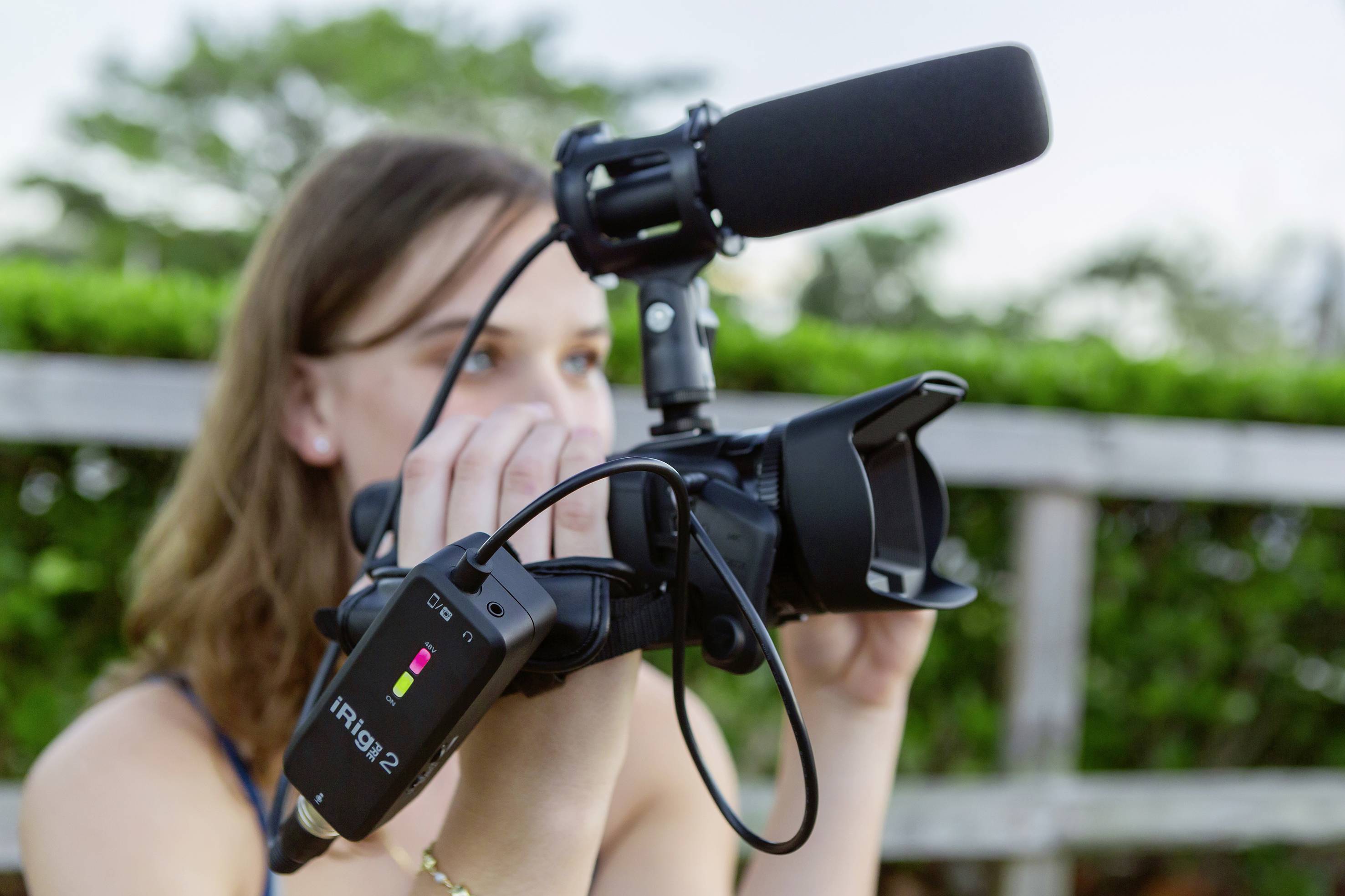 Eine Person filmt im Freien mit einer Videokamera, ausgestattet mit einem Mikrofon und einem iRig2-Audiointerface. Im Hintergrund sind Bäume sichtbar.