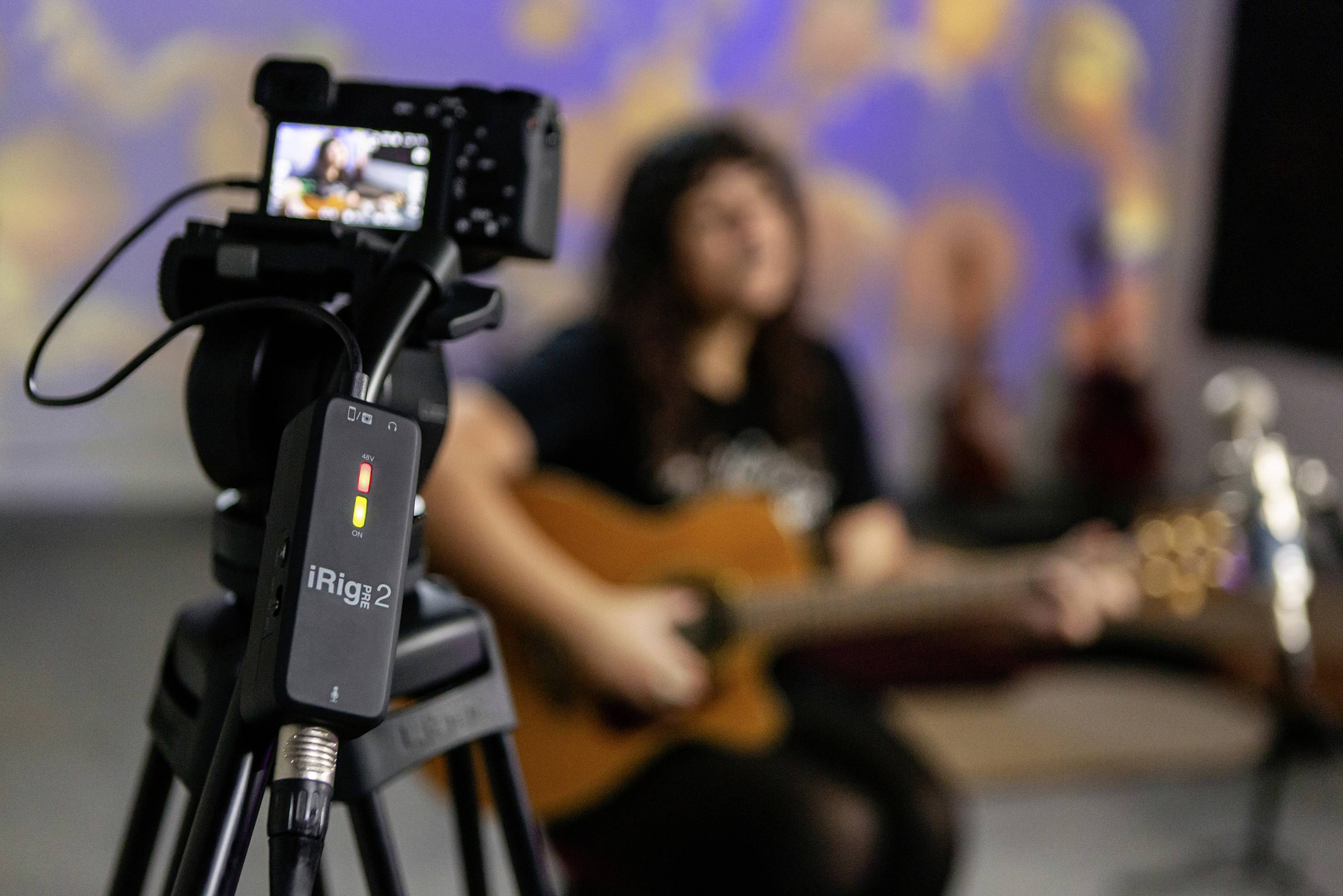 Eine Kamera im Vordergrund filmt eine sitzende Person, die Gitarre spielt. Im Hintergrund ist die Person unscharf dargestellt.