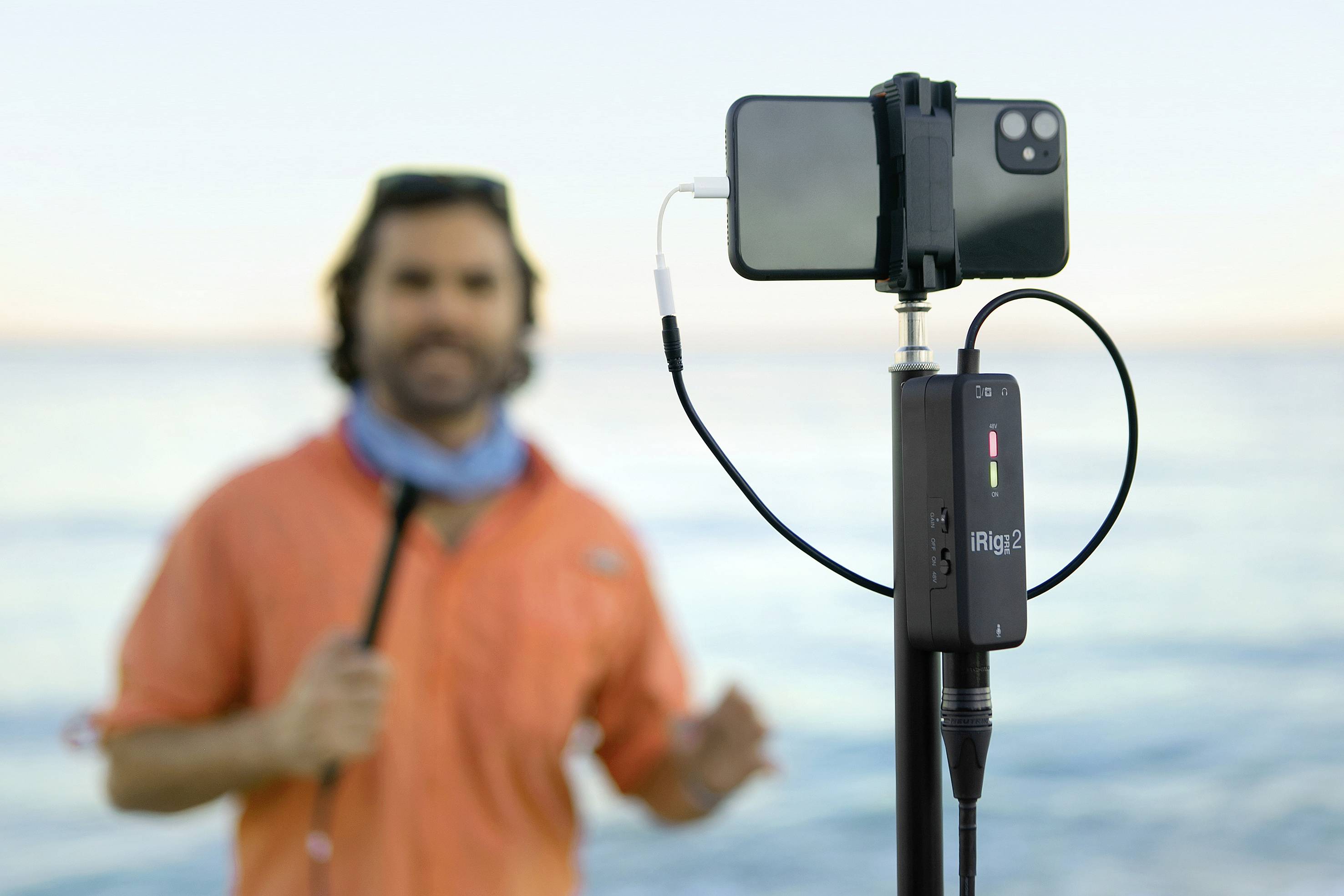 Ein Mann mit oranger Kleidung steht am Meer und wird mit einem Smartphone auf einem Stativ gefilmt. Im Vordergrund ist ein Audio-Interface zu sehen.