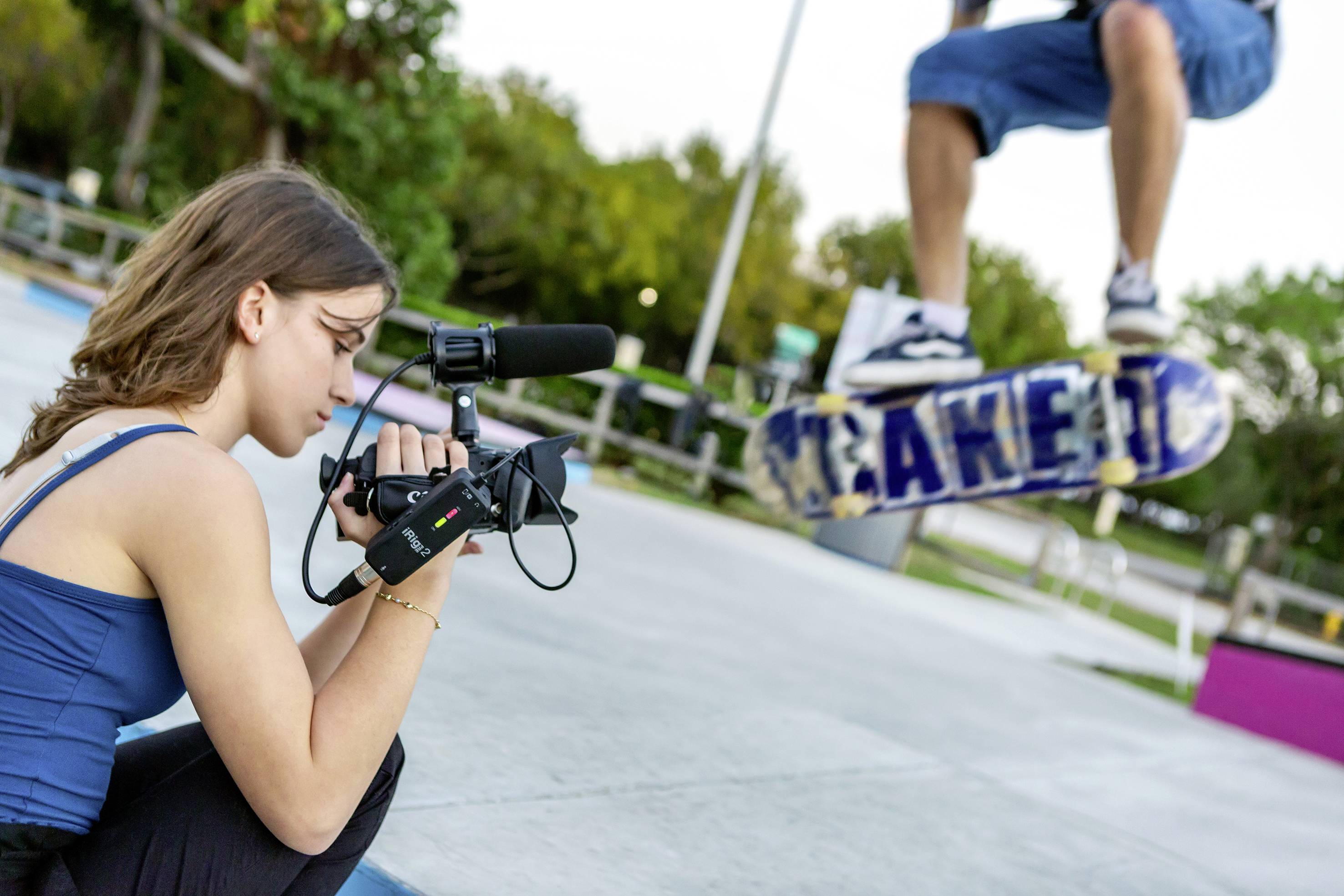 Eine junge Frau filmt mit einer Videokamera einen Skateboarder, der in der Luft springt. Die Umgebung ist ein Skatepark.