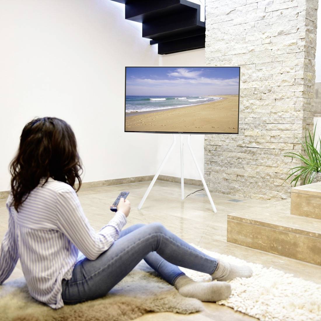 Eine Frau sitzt auf einem Teppich in einem Wohnzimmer und schaut Fernsehen. Der Bildschirm zeigt eine Strandlandschaft. Sie hält eine Fernbedienung in der Hand.