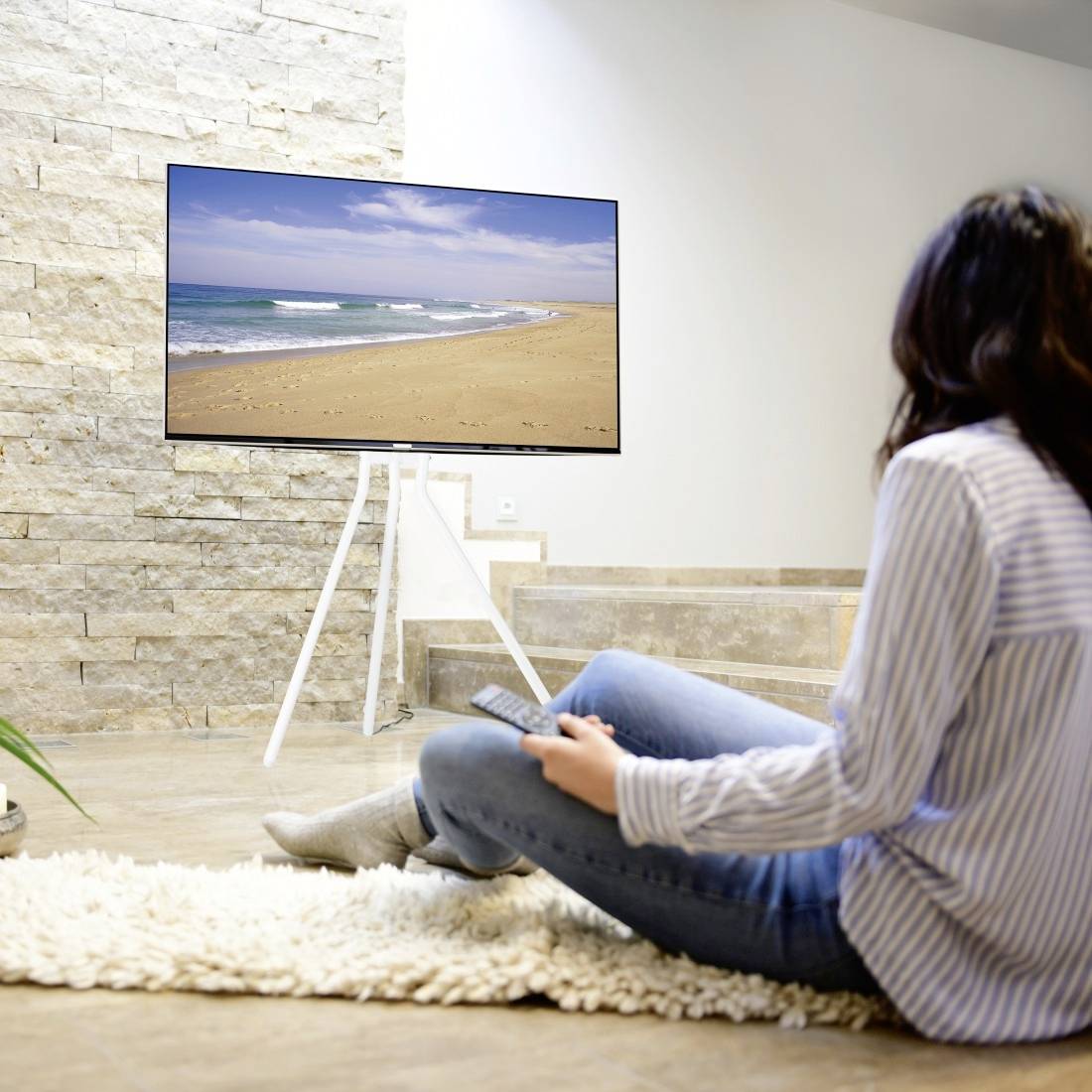 Eine Person sitzt in einem modernen Wohnzimmer und schaut auf einen großen Flachbildfernseher, auf dem ein Strandbild gezeigt wird.