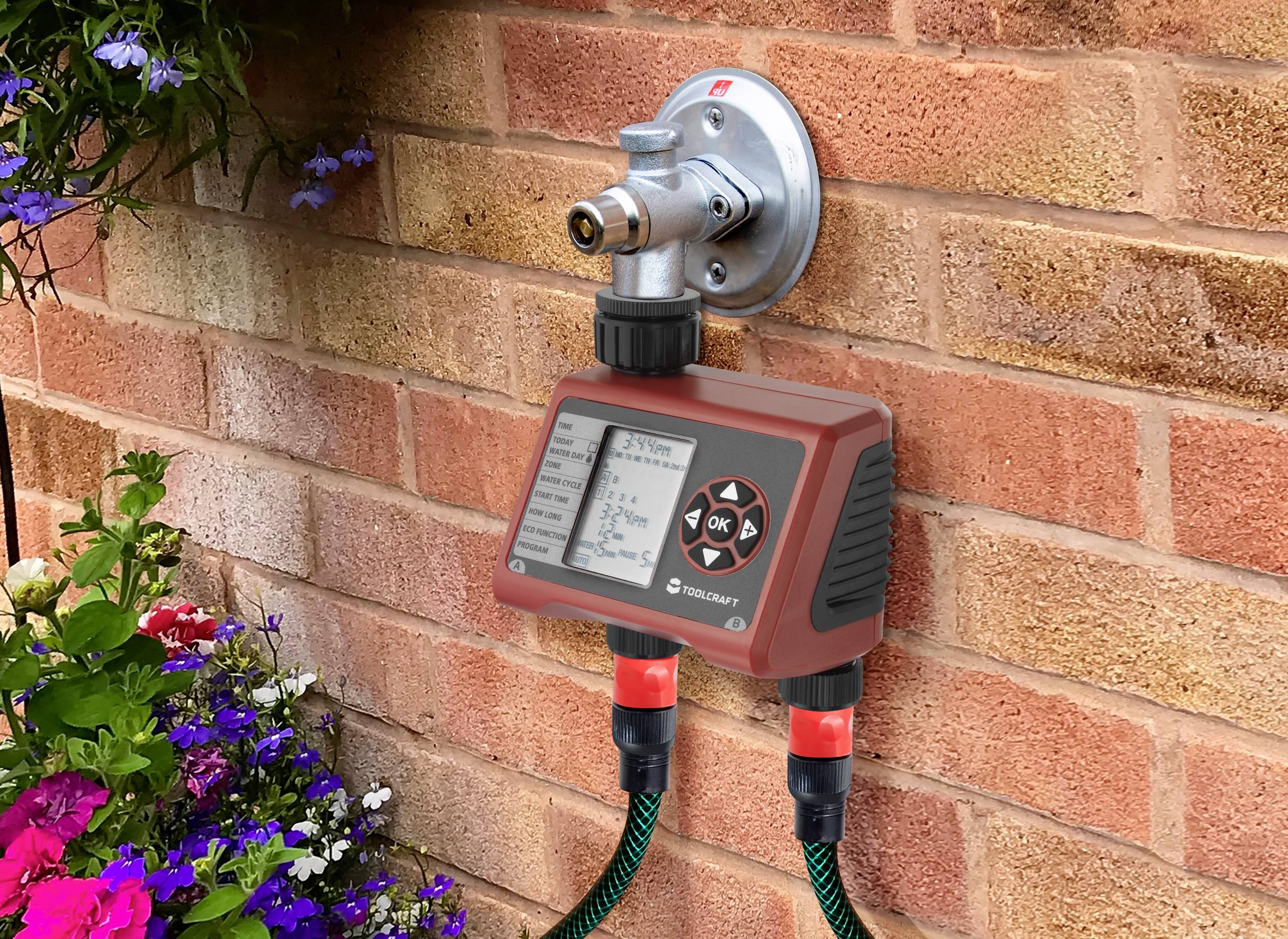 Wasserhahn mit digitalem Bewässerungssteuergerät an einer Ziegelwand. Unterhalb befinden sich zwei Gartenschläuche und Blumenbeet.