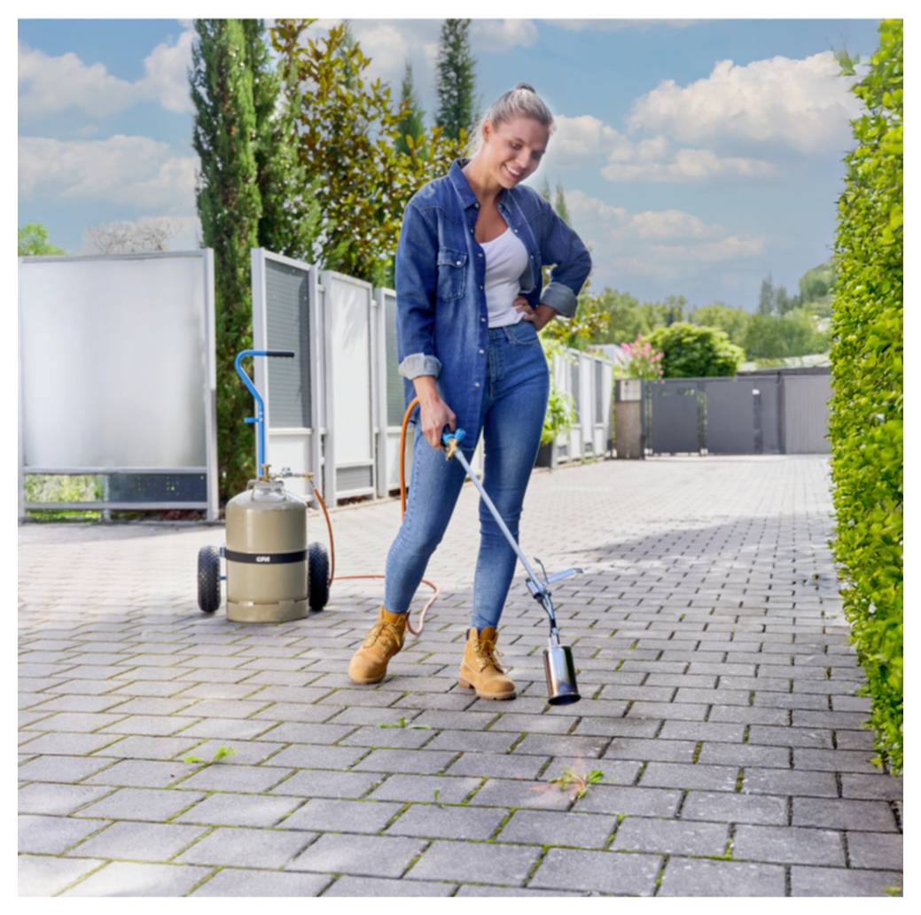 Frau, die einen Unkrautvernichter auf Terrassenplatten verwendet, mit einem Propantank auf einem Wagen mit Rädern hinter ihr.
