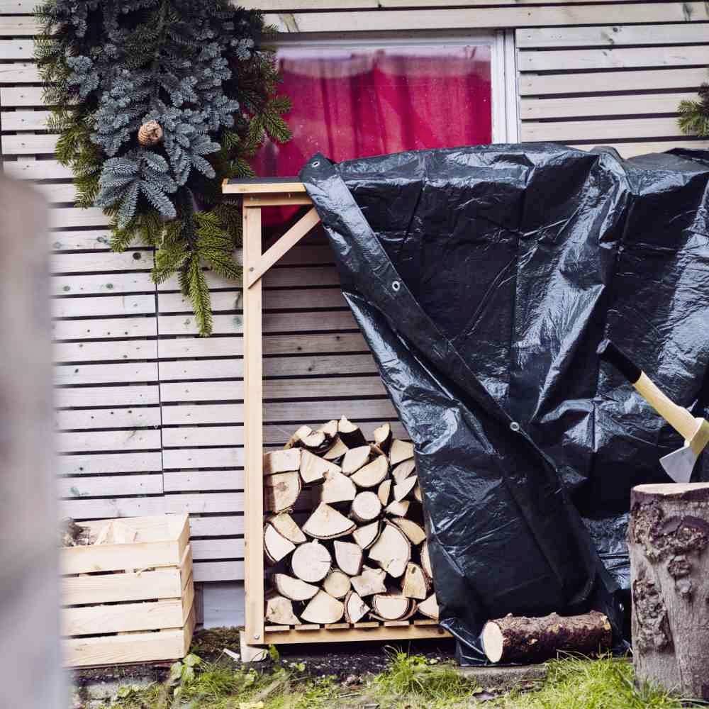Ein Stapel Holz liegt unter einer dunkelgrünen Plane neben einer Holzkiste mit weiteren Holzscheiten. Ein Kranz hängt an der Wand.