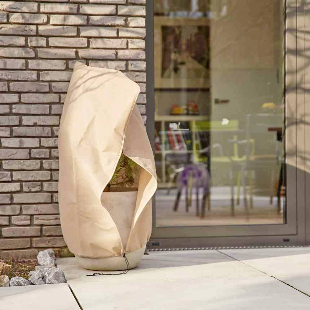 Ein eingepackter Topf mit Pflanze steht auf einer Terrasse neben einer Glasfassade vor einer Ziegelsteinwand.