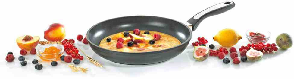 Eine Bratpfanne mit einem Pfannkuchen, belegt mit Himbeeren, Blaubeeren und Apfelscheiben. Umgeben von Obst wie Zitrone und Kiwi.