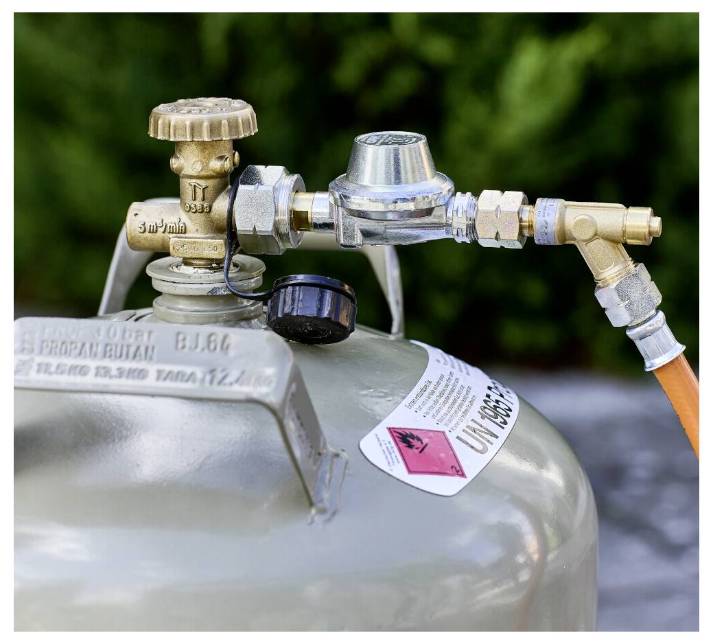 Nahaufnahme eines Gasflaschenventils und eines Regulators, die mit einem orangefarbenen Schlauch verbunden sind und auf eine Druckgasanlage hindeuten.