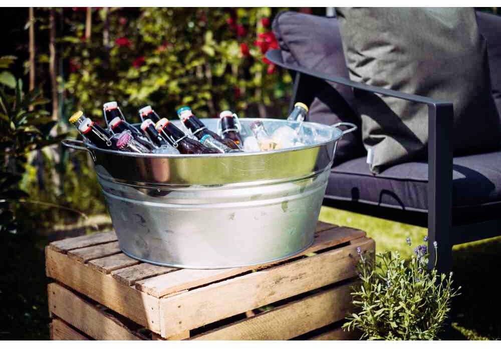 Metallwanne mit Eis und Flaschen auf Holzkiste im Garten, neben Stuhl mit Kissen.