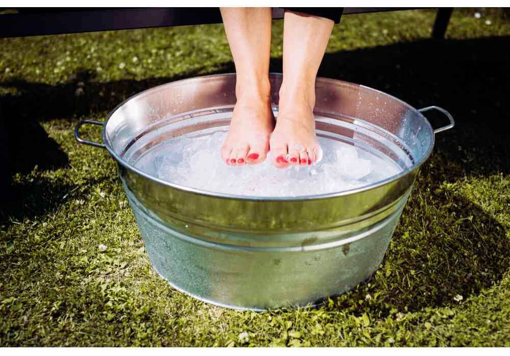 Person taucht ihre Füße in einen Eimer mit Eiswasser, steht im Freien auf Gras. Entspannungs- oder Abkühlungsaktion.