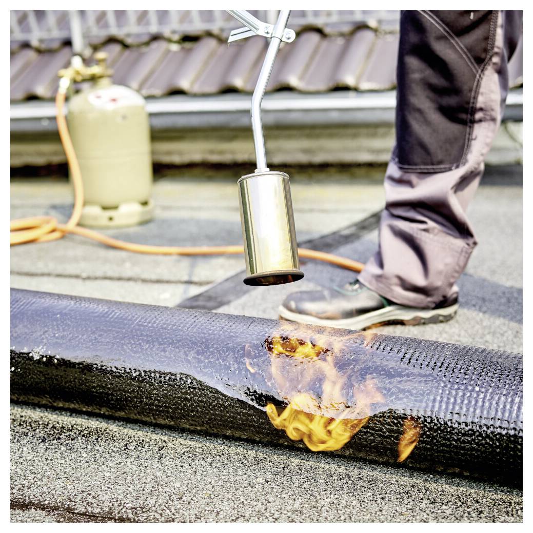 Arbeiter, der eine Lötlampe verwendet, um die Dachdeckungsmembran auf einem Flachdach zu erwärmen und zu versiegeln, wobei Flammen entlang der Naht sichtbar sind.