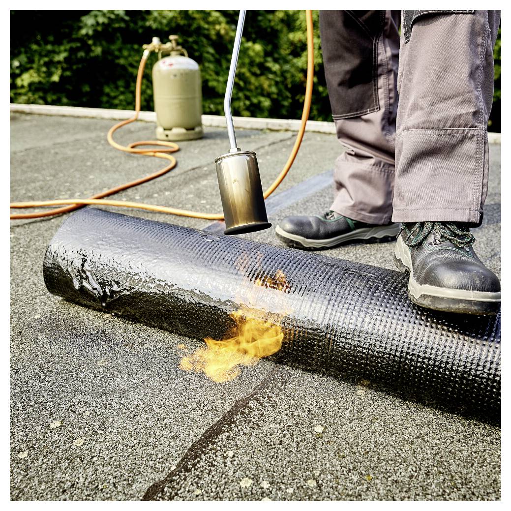 Ein Arbeiter nutzt einen Brenner, um eine aufgerollte Dachpappe auf einem Flachdach zu erhitzen und zu versiegeln.