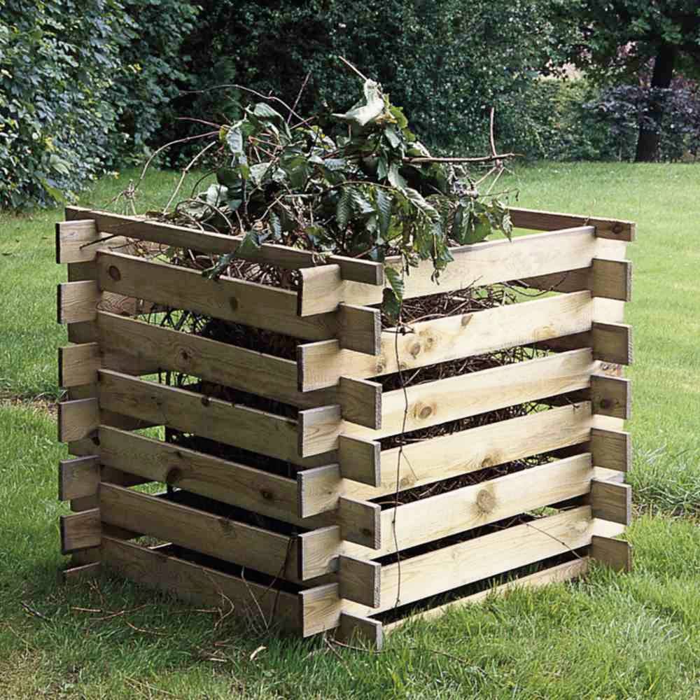 Holzkomposter auf Rasen, gefüllt mit Gartenabfällen wie Zweigen und Blättern, umgeben von Bäumen im Hintergrund.