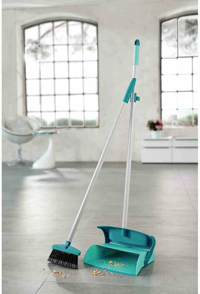 Besen und Kehrschaufel stehen auf einem Boden mit Krümeln. Im Hintergrund sind große Fenster und ein Stuhl sichtbar.