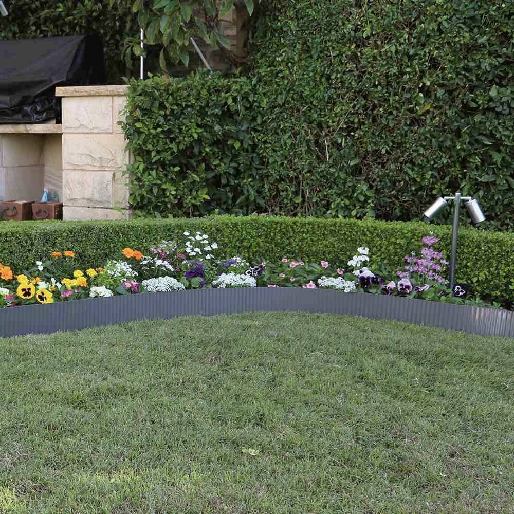 Ein gepflegter Garten mit einer Hecke und einem Blumenbeet voller bunter Blumen. Links ein gemauerter Grill und grüner Rasen.