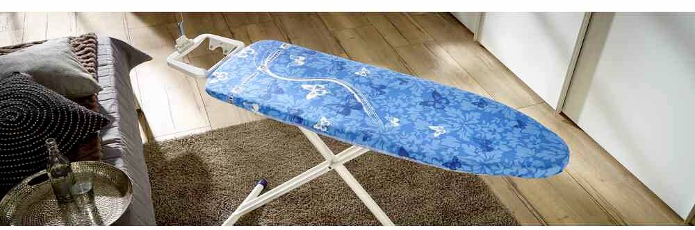 Bügelbrett mit blauem Blumenbezug steht auf einem Teppich in einem Wohnraum.