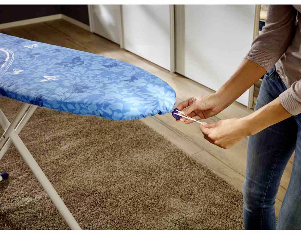 Eine Person zieht an einem blauen Bügelbrettbezug mit Blumenmuster, das in einem Raum auf einem braunen Teppich steht.