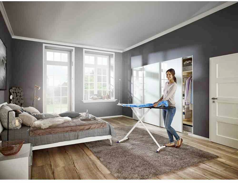 Eine Person bügelt ein Hemd auf einem Bügelbrett in einem modern eingerichteten Schlafzimmer mit grauen Wänden und Holzfußboden.