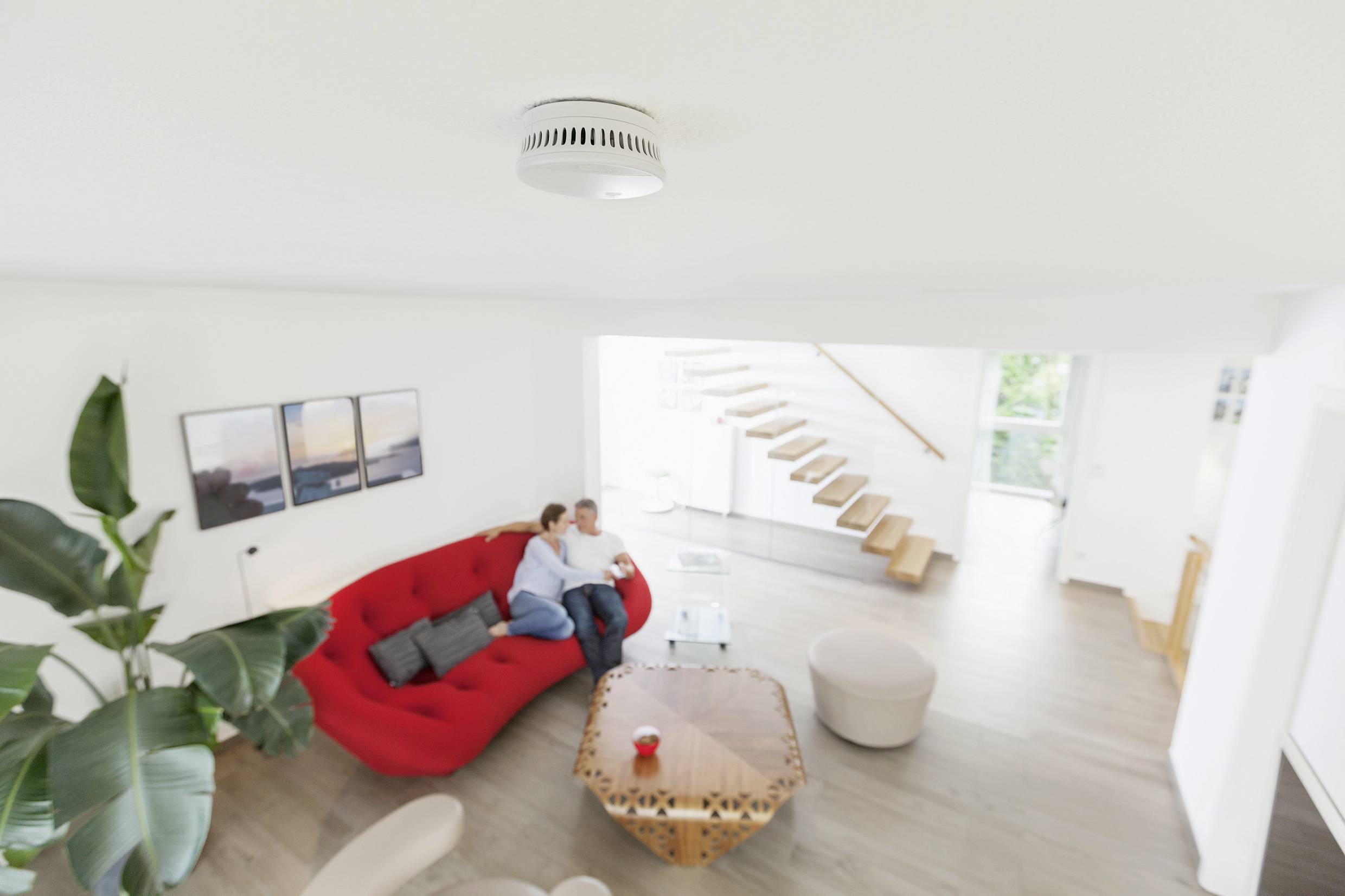 Ein Paar sitzt entspannt auf einem roten Sofa in einem modernen Wohnzimmer. Auf einem Tisch in der Mitte steht eine rote Tasse.