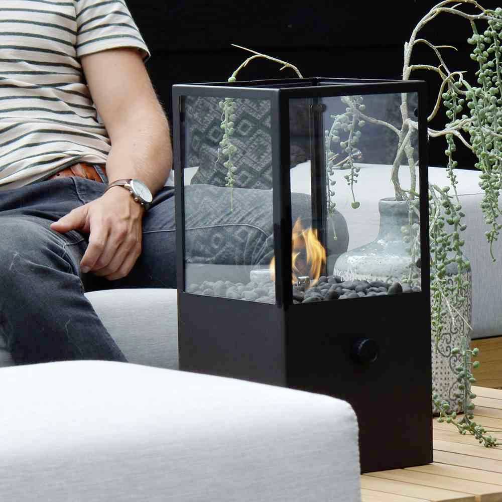 Eine Person in gestreiftem Hemd sitzt neben einem modernen, gläsernen Kaminofen. Im Hintergrund befinden sich Pflanzen und ein Kissen.