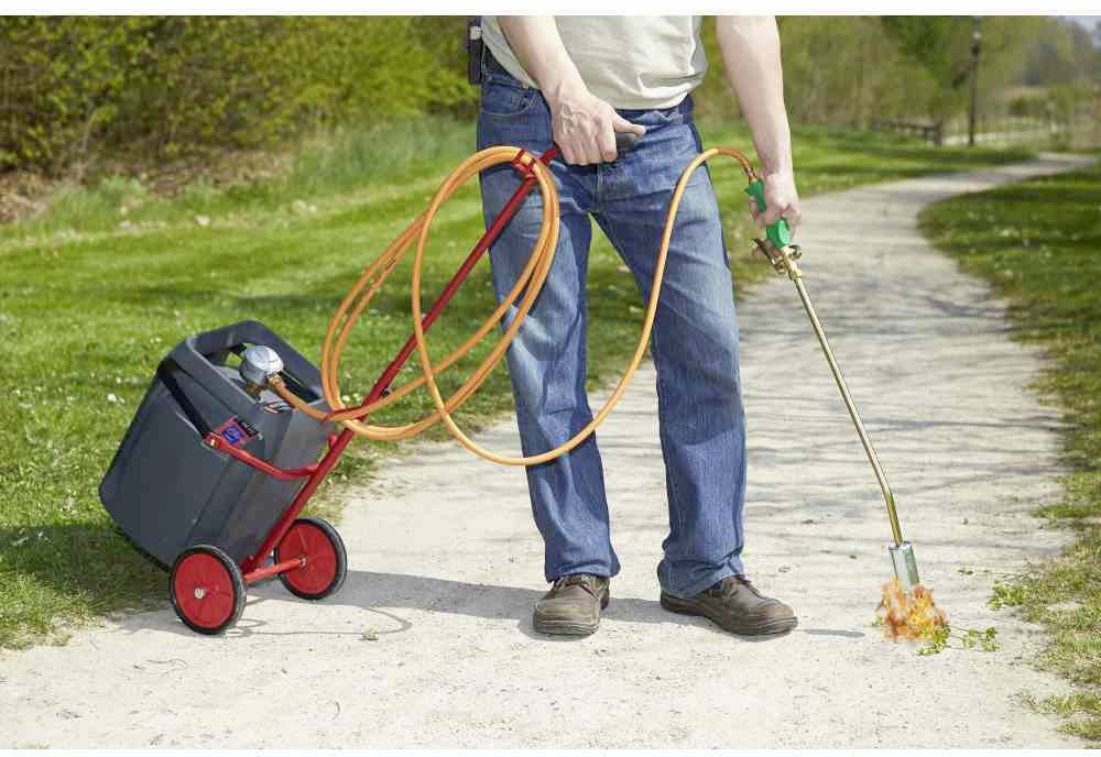 Eine Person verwendet einen Brenner mit Gasflasche auf einem Weg, um Unkraut zu beseitigen. Der Brenner ist mit einem Schlauch verbunden.