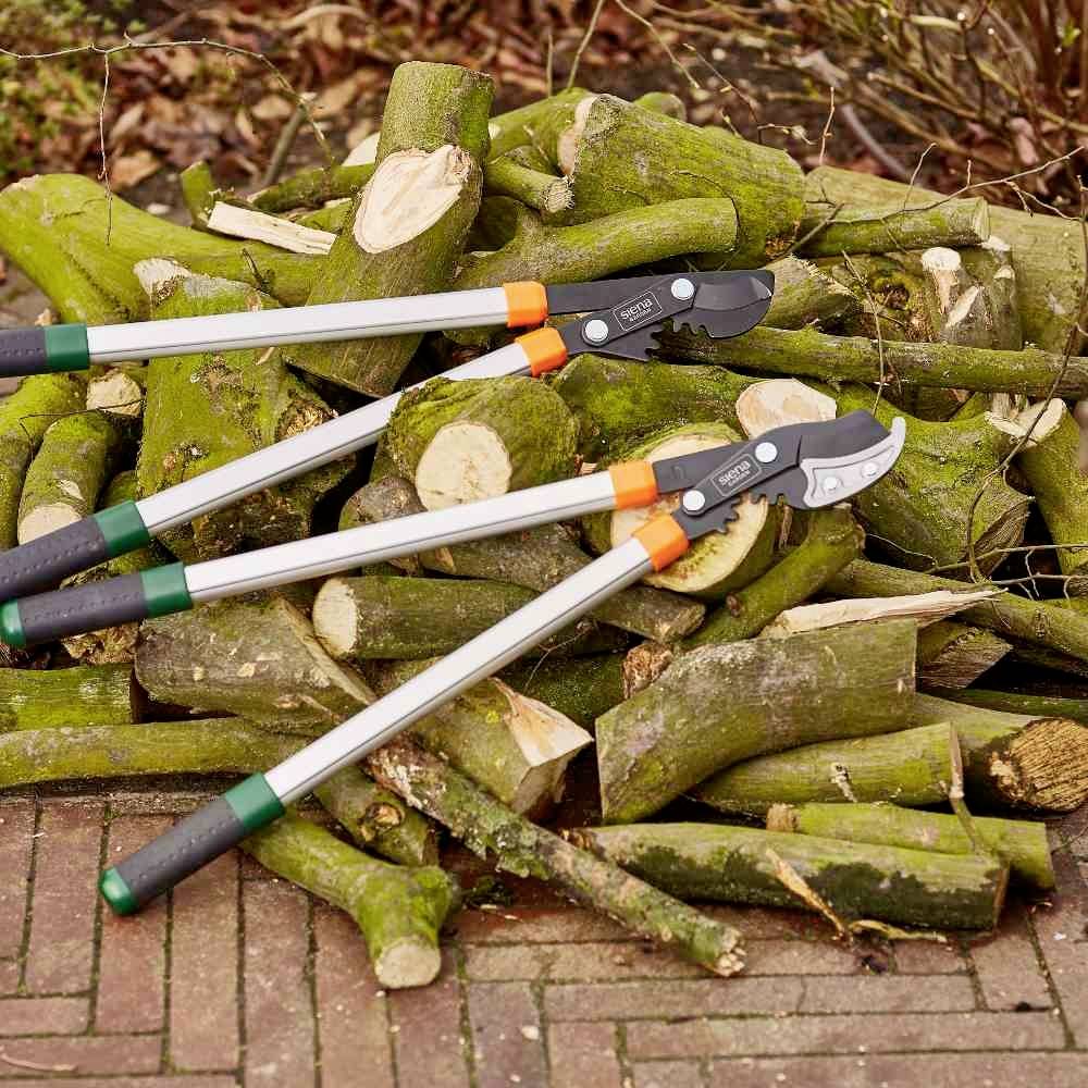 Ein Stapel geschnittener Holzscheite liegt auf dem Boden, umgeben von zwei Astscheren mit langen Griffen, bereit zum Beschneiden.