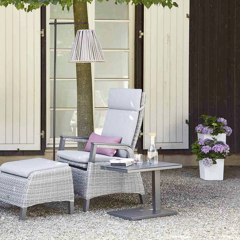 Ein gemütlicher Gartenstuhl und Hocker aus Rattan stehen unter einem Baum. Ein kleiner Tisch mit Getränk und Blumen verleiht eine einladende Atmosphäre.