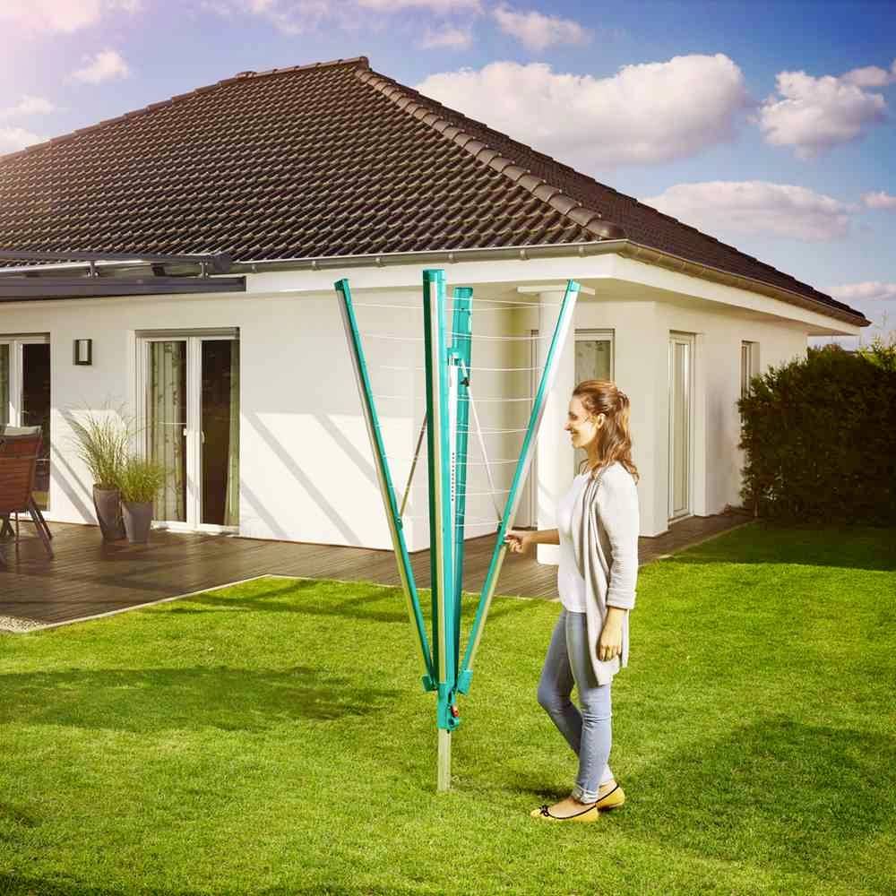 Eine Frau steht auf einem sonnigen Rasen vor einem modernen Einfamilienhaus und klappt einen grünen, zusammengefalteten Wäscheschirm auf.