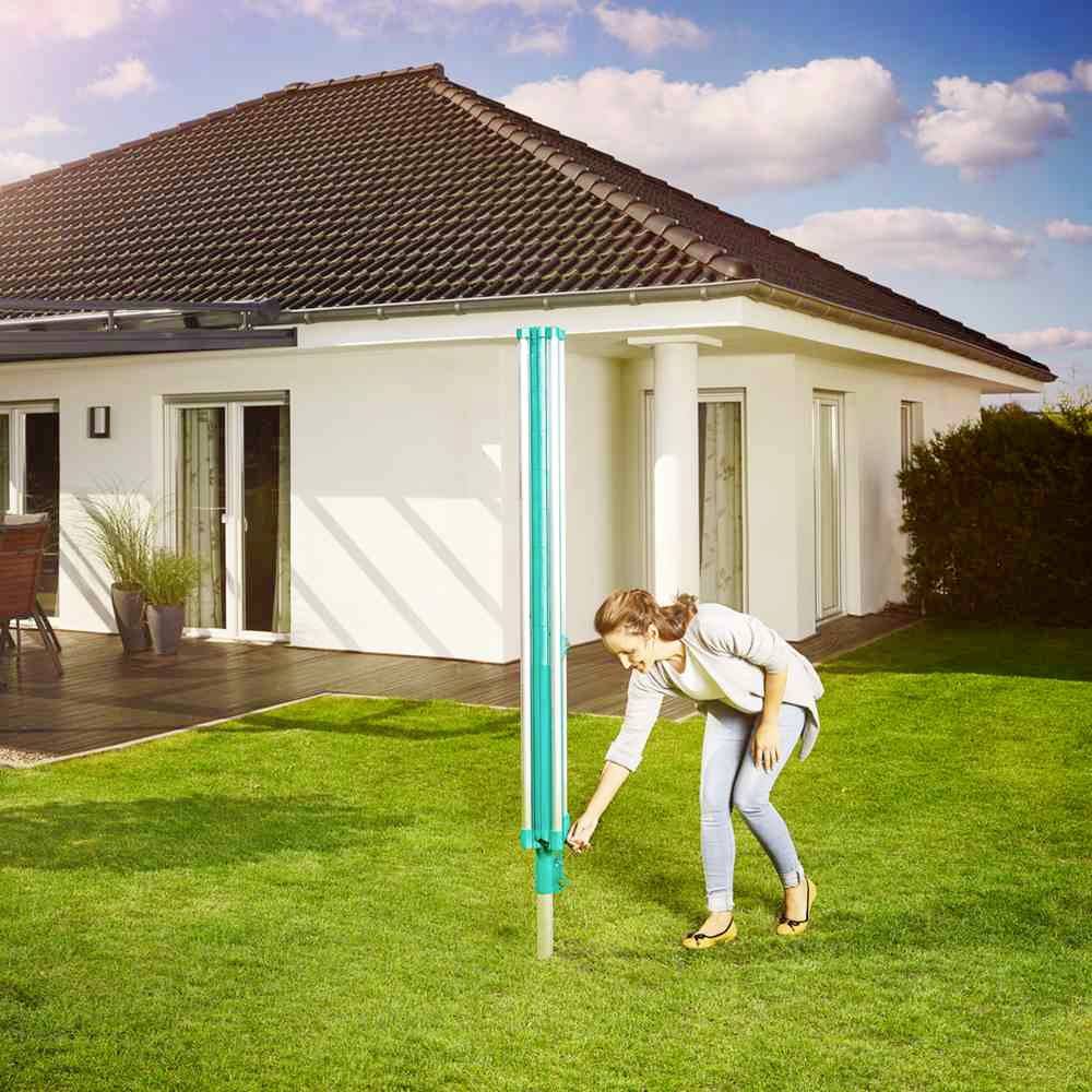 Eine Frau installiert einen blauen Wäscheschirm in einem saftig grünen Garten vor einem modernen Haus mit dunklem Dach und Terrasse.
