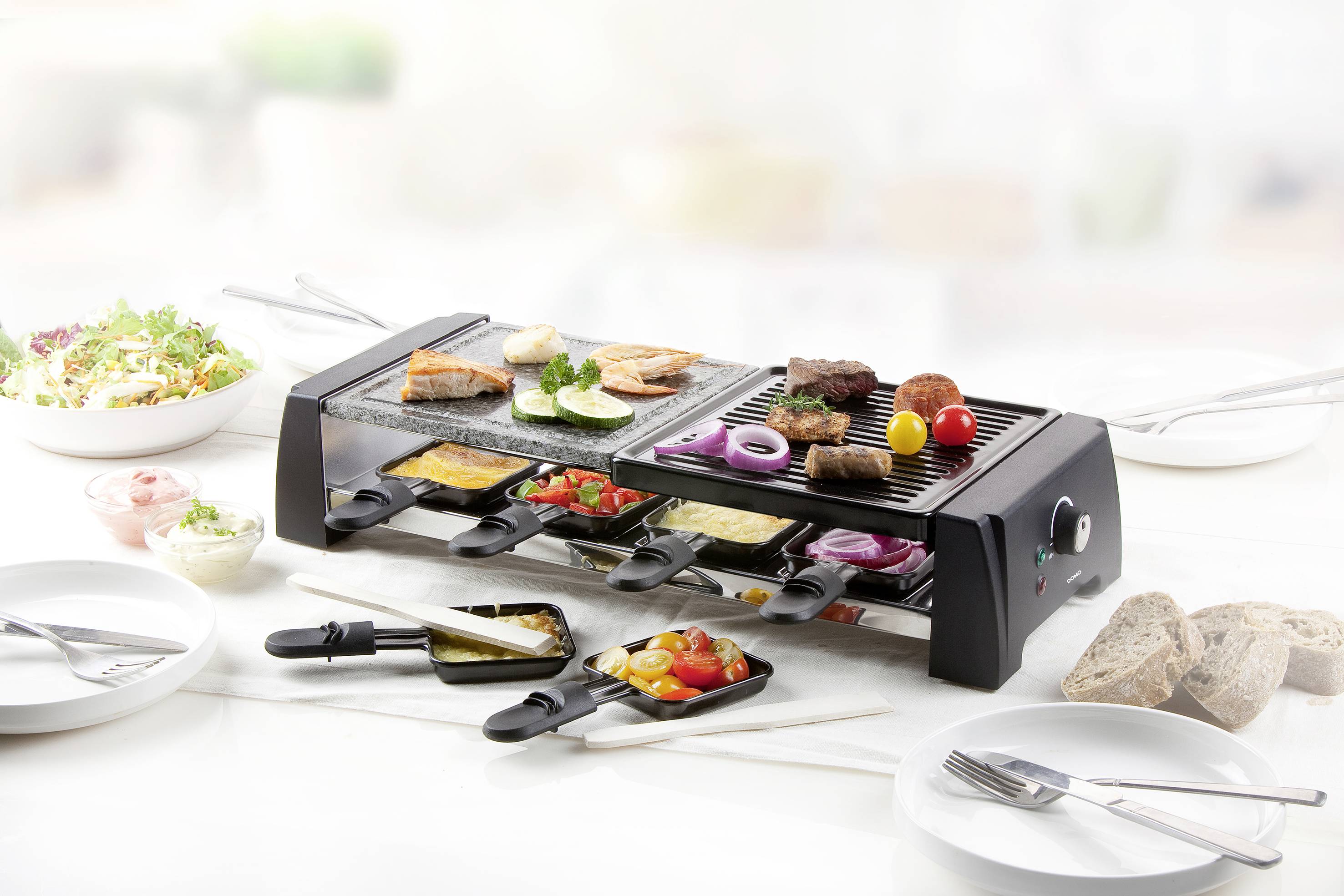 Ein Raclette-Grill mit verschiedenen Zutaten wie Fleisch, Fisch, Gemüse und Käse, umgeben von Beilagen wie Salat und Brot.