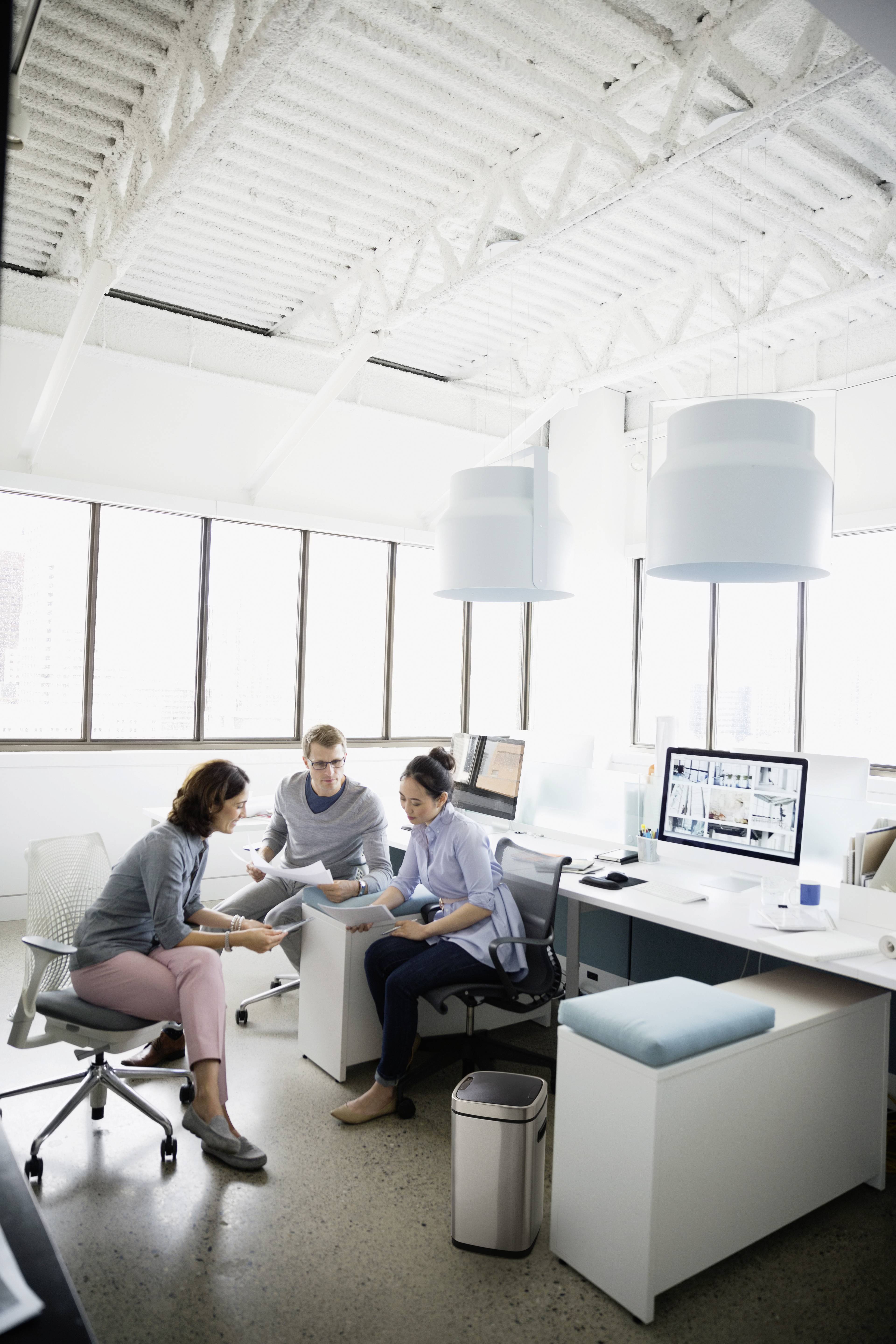 Drei Personen in einem modernen Büro arbeiten zusammen. Sie sitzen an Schreibtischen mit Computern, umgeben von großen Fenstern.