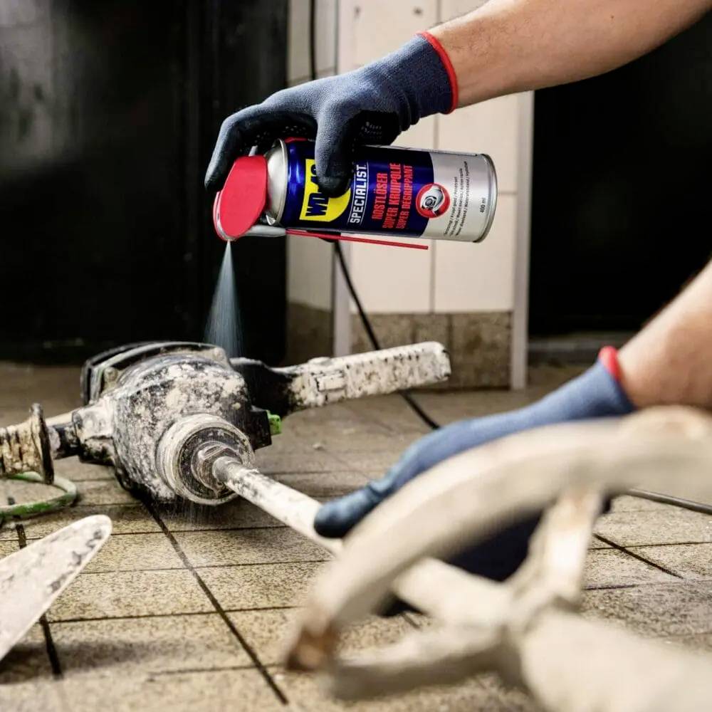 Eine Person reinigt ein Metallwerkzeug mit einem Schmiermittel, das aus einer Spraydose aufgetragen wird. Sie trägt blaue Handschuhe.