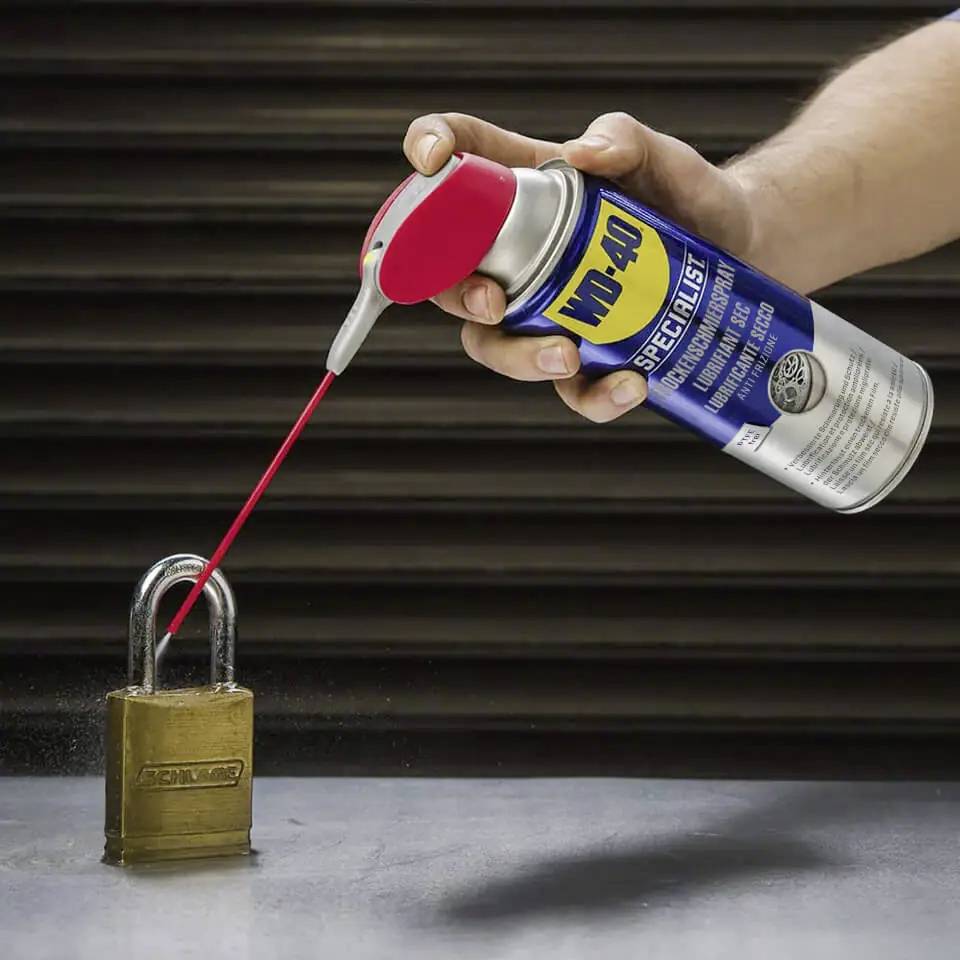 Eine Hand sprüht WD-40 mit einer roten Sprühröhrchen auf ein Vorhängeschloss und deutet damit auf die Verwendung des Produkts zum Schmieren und Schützen von Schlössern hin.