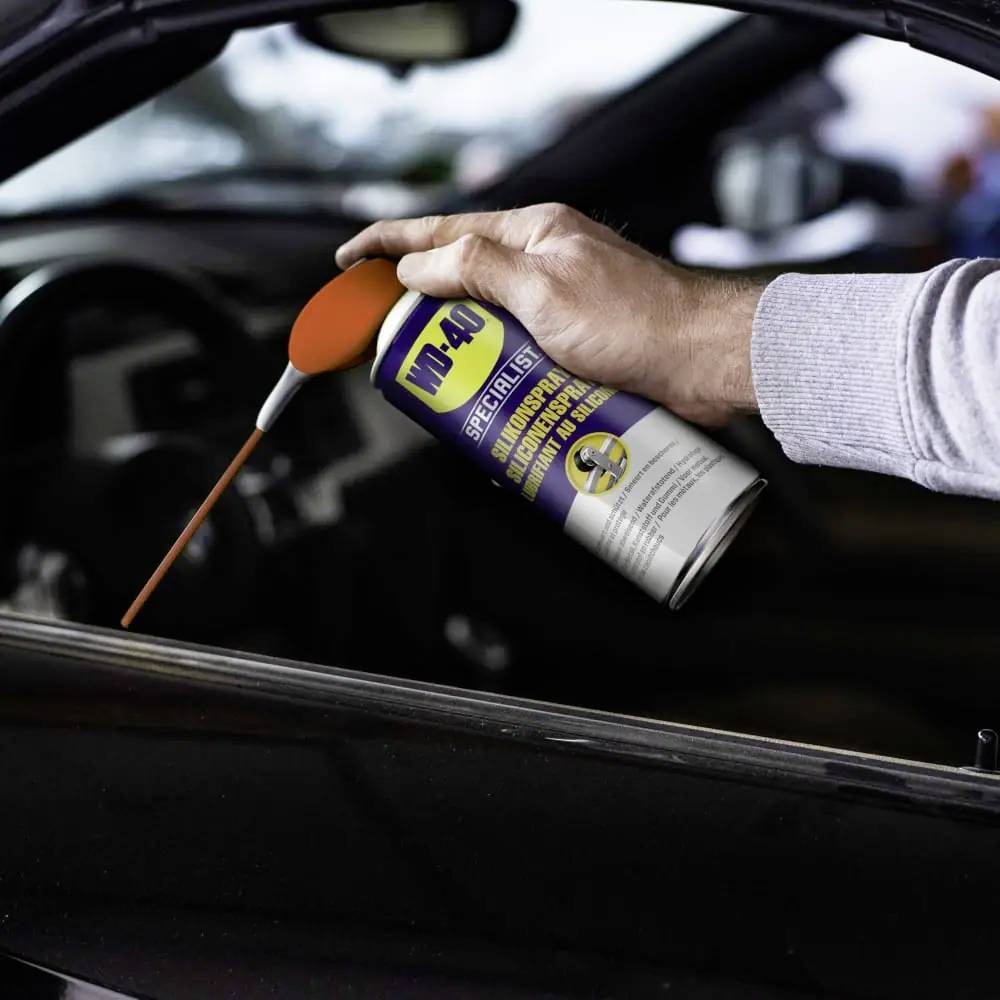 Eine Person hält eine WD-40-Dose und sprüht Schmiermittel auf einen Autoteil. Die Dose hat einen orangefarbenen Sprühkopf.