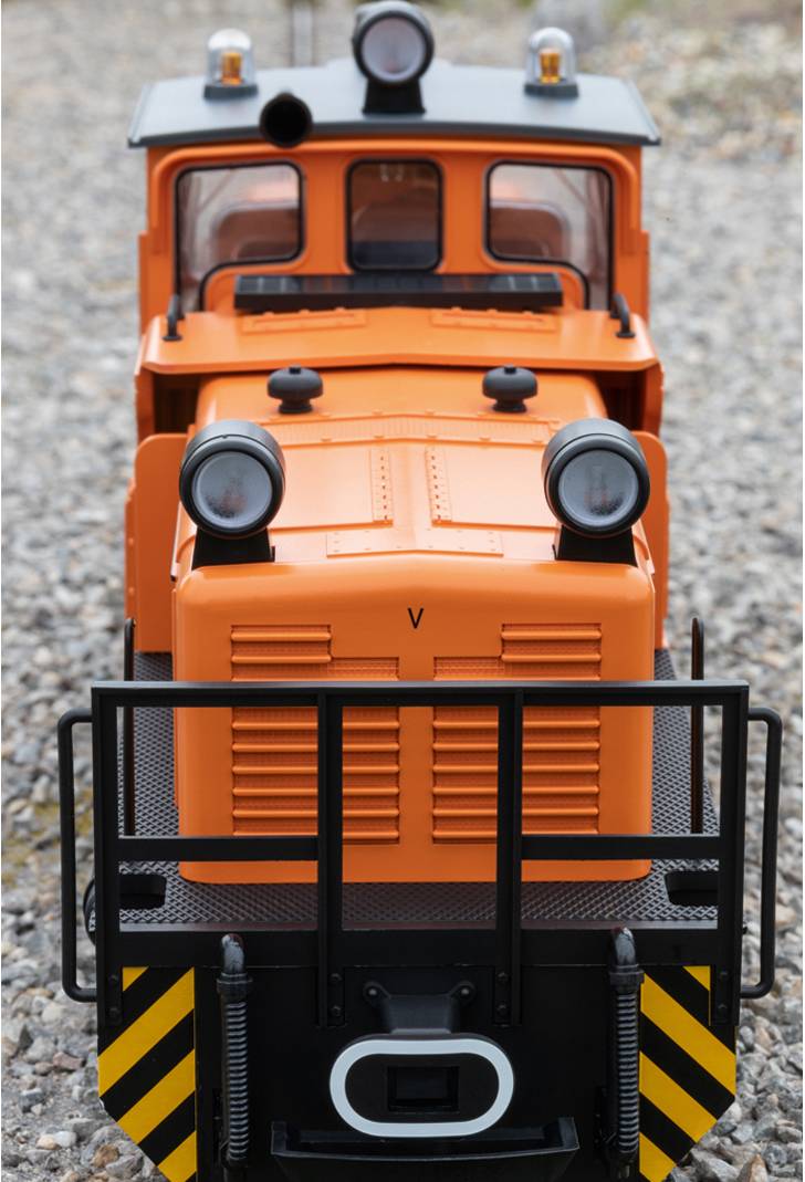 Kleine orangene Lokomotive auf einem Kiesweg, frontal fotografiert. Sie besitzt zwei große Scheinwerfer und eine schwarze Schutzvorrichtung vorne.