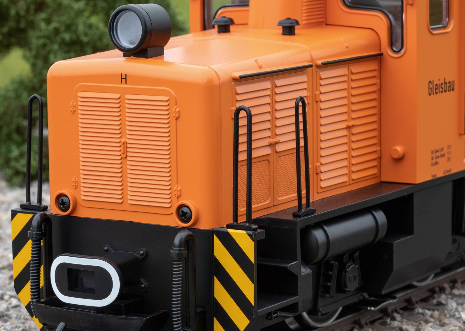 Orange Lokomotive mit schwarzen Streifen, Aufschrift 'Gleisbau', detaillierte Vorderseite, Umgebung mit grüner Vegetation sichtbar.