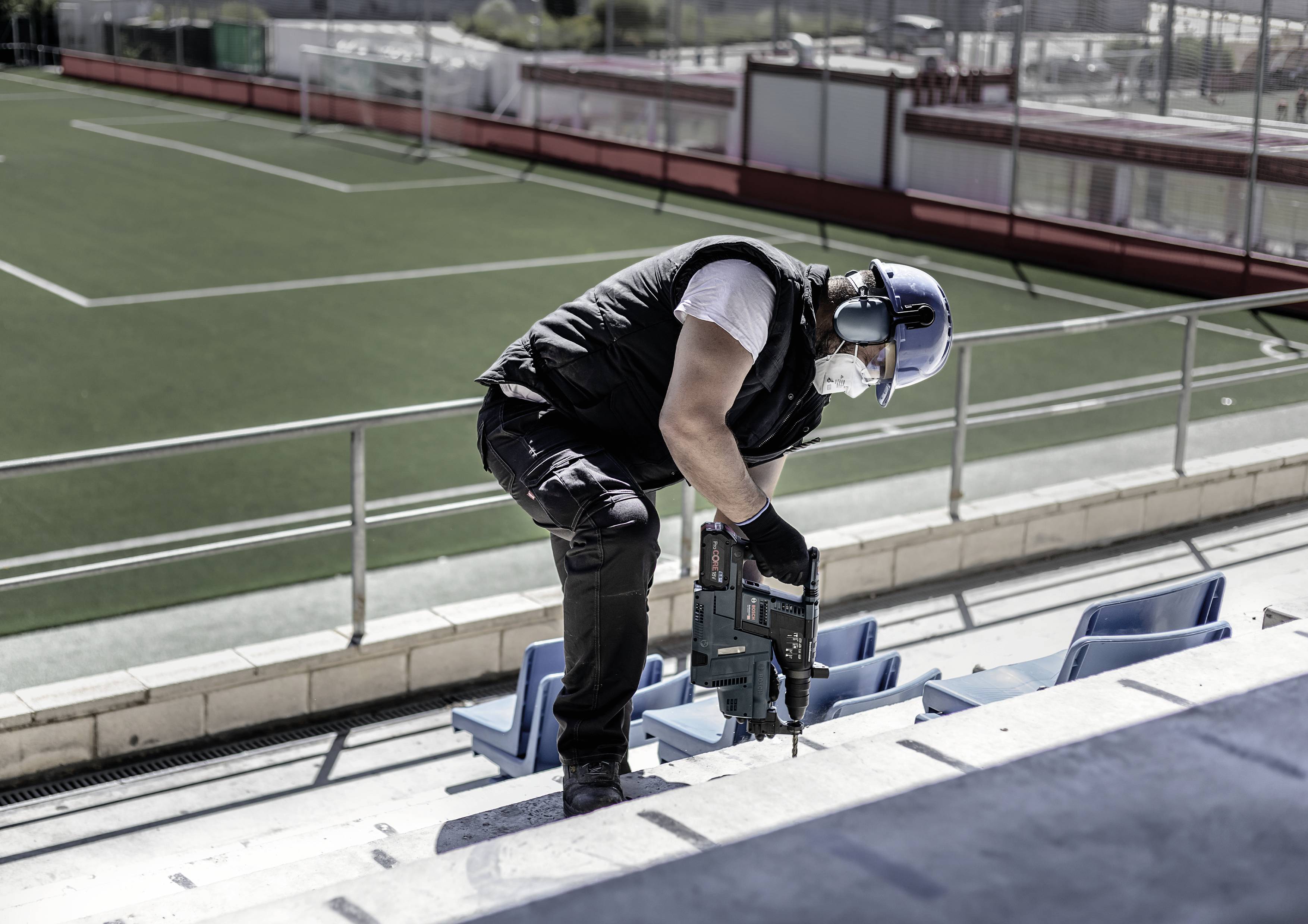 Eine Person arbeitet mit einem elektrischen Werkzeug auf den Stufen eines Stadions, im Hintergrund ist ein Fußballfeld sichtbar.