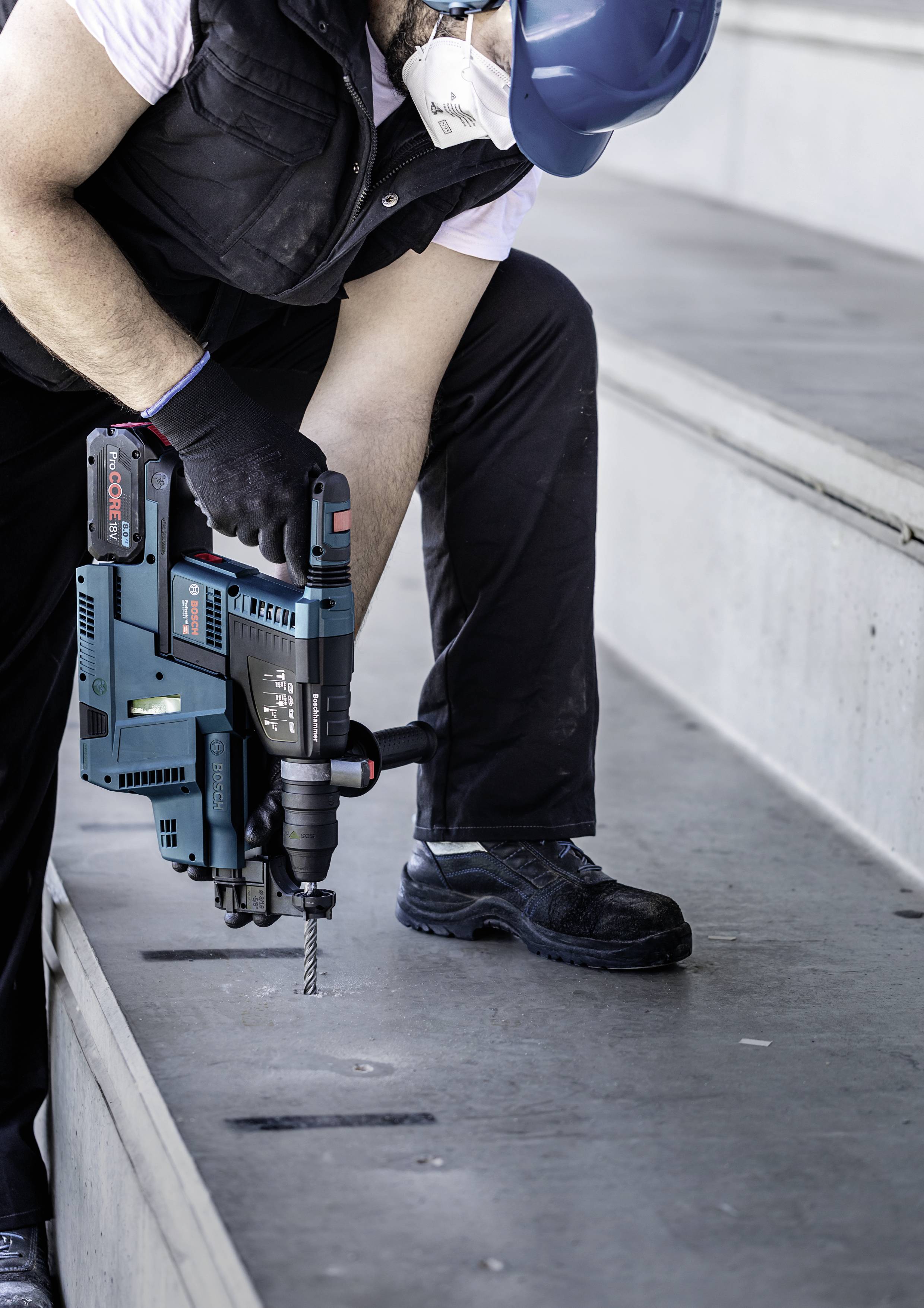 Person, die einen blauen Bauhelm und eine Maske trägt, verwendet einen elektrischen Bohrer auf einer Betonstufe.