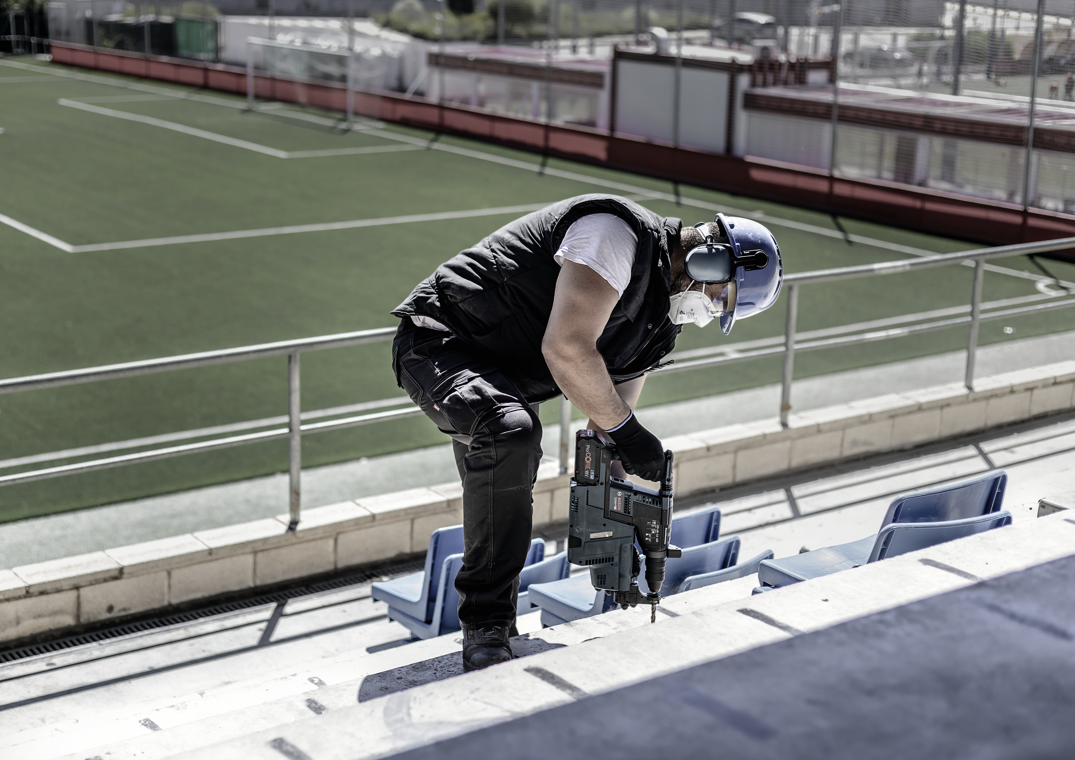 Arbeiter mit Schutzhelm und Gehörschutz führt Bauarbeiten auf Stufen eines leeren Sportstadions durch.