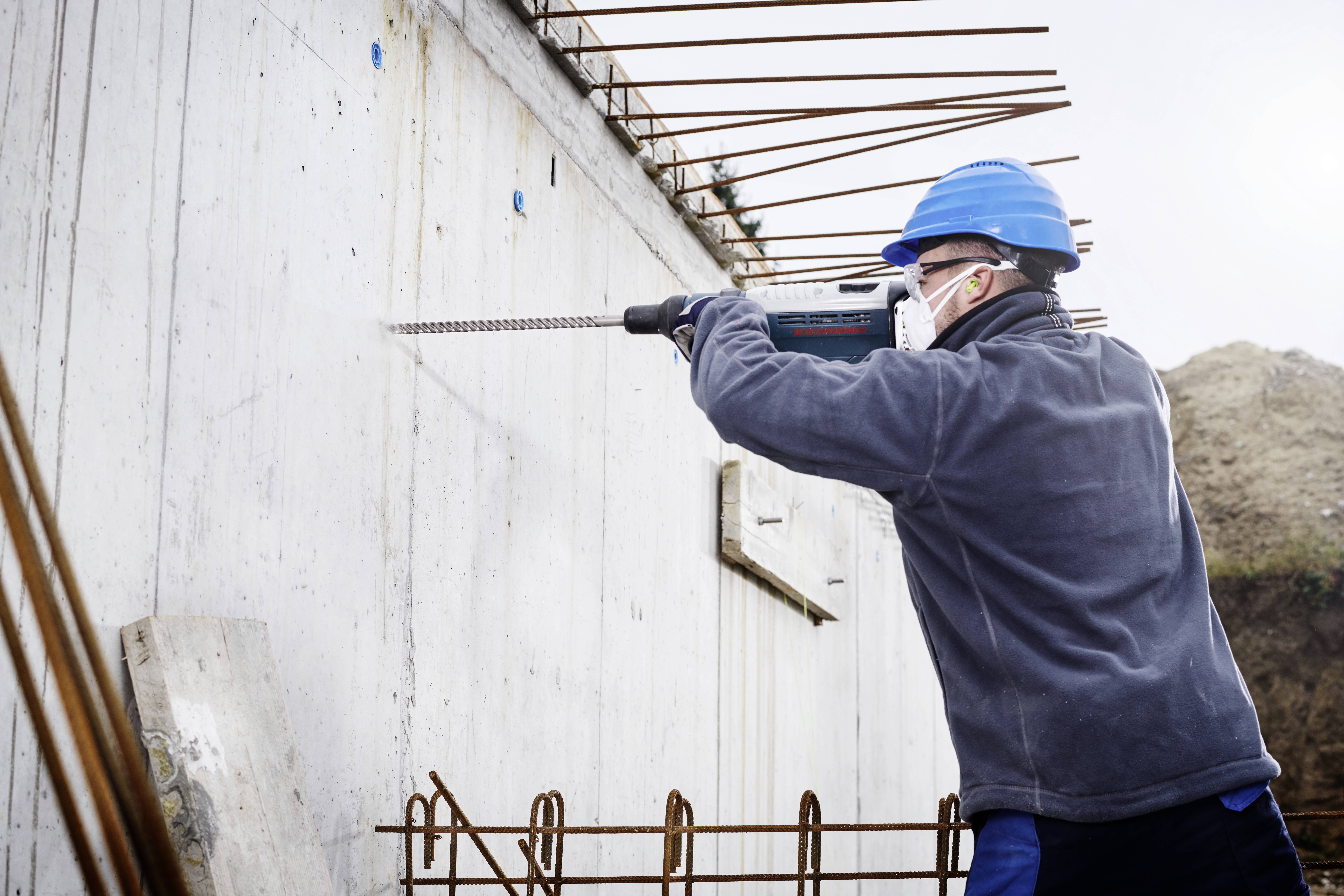 Eine Person in Schutzkleidung und blauem Helm verwendet eine Bohrmaschine, um Löcher in eine Betonwand auf einer Baustelle zu bohren.
