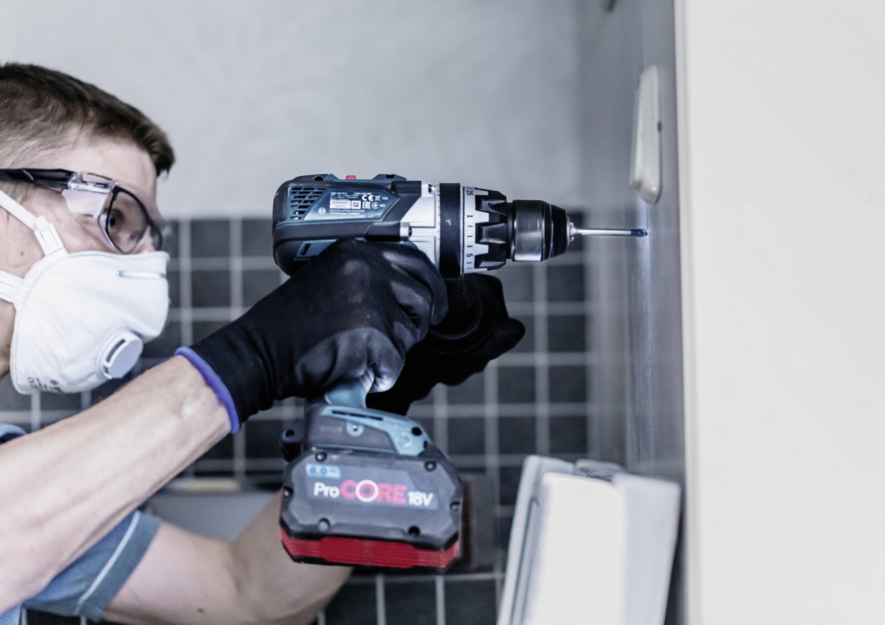 Ein Mann mit Schutzbrille und Maske bohrt mit einer elektrischen Bohrmaschine ein Loch in eine geflieste Wand.