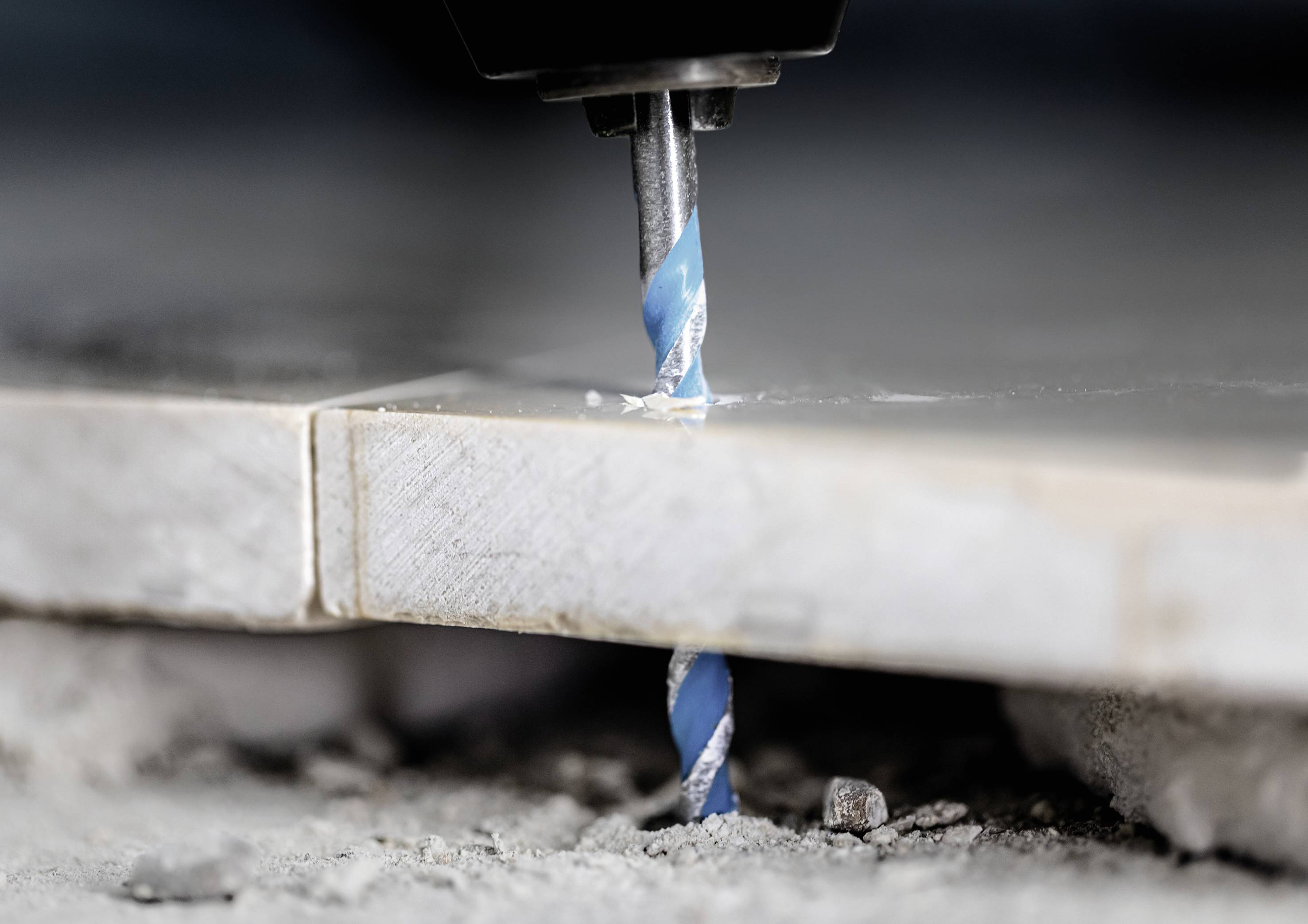 Eine Bohrmaschine durchbohrt eine Fliese. Der Bohrer hat eine blaue Spirale. Staub und Fliesenreste sind auf dem Boden sichtbar.