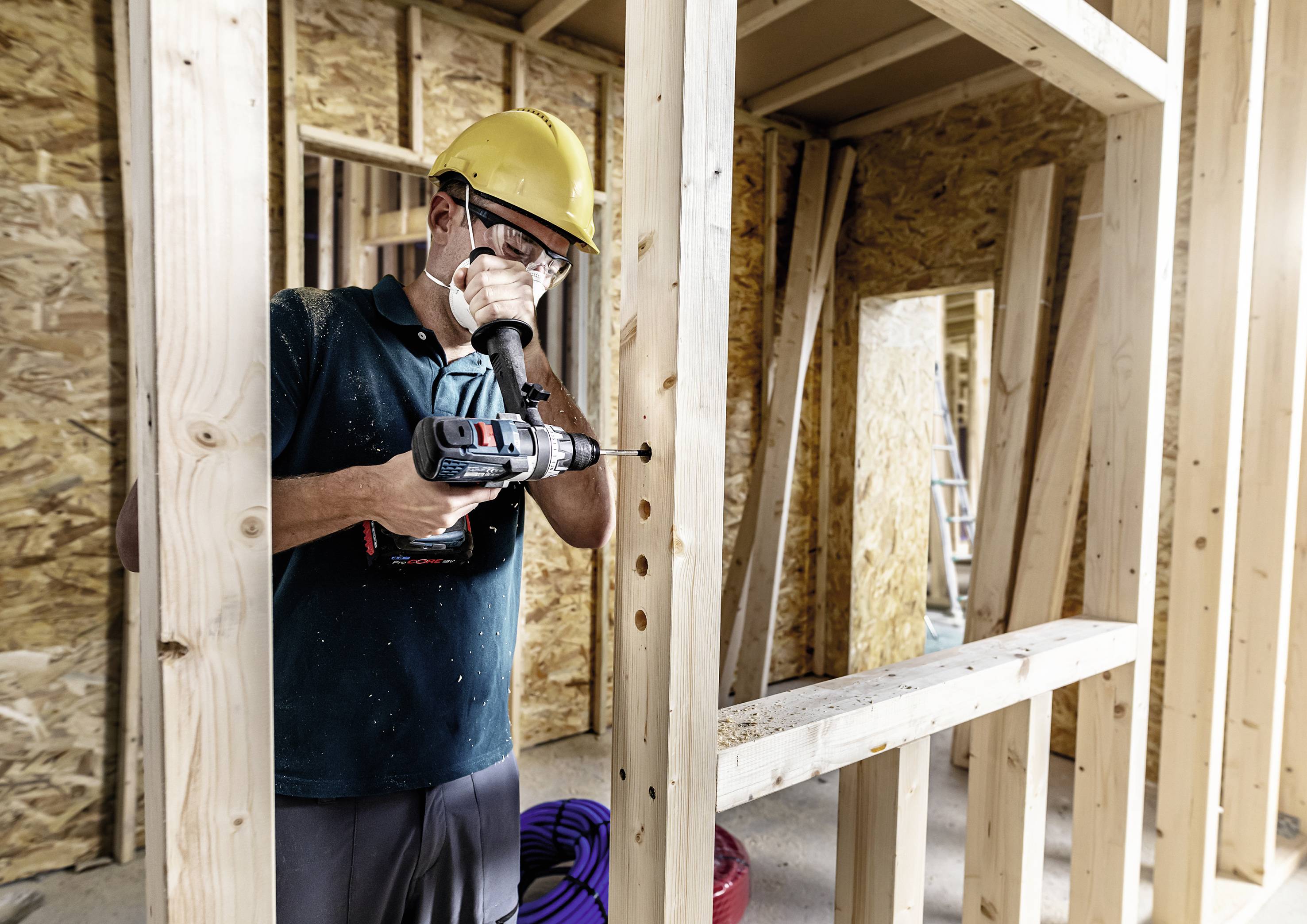 Eine Person in Schutzkleidung benutzt eine Bohrmaschine, um Holzrahmen in einem im Bau befindlichen Gebäude zu verschrauben.