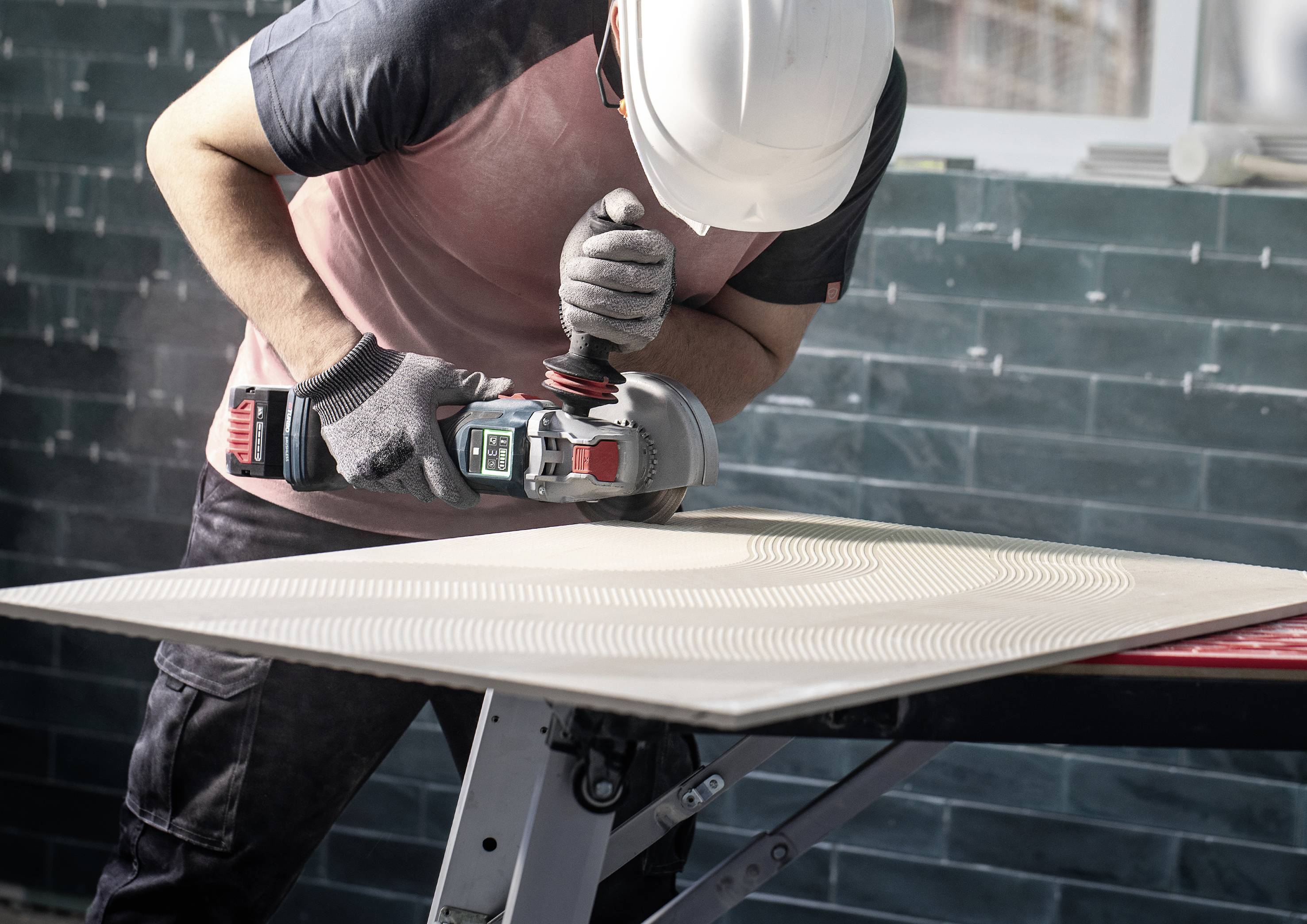 Ein Arbeiter mit Helm schleift eine große Holzplatte in einer Werkstatt. Er nutzt dazu einen elektrischen Winkelschleifer.