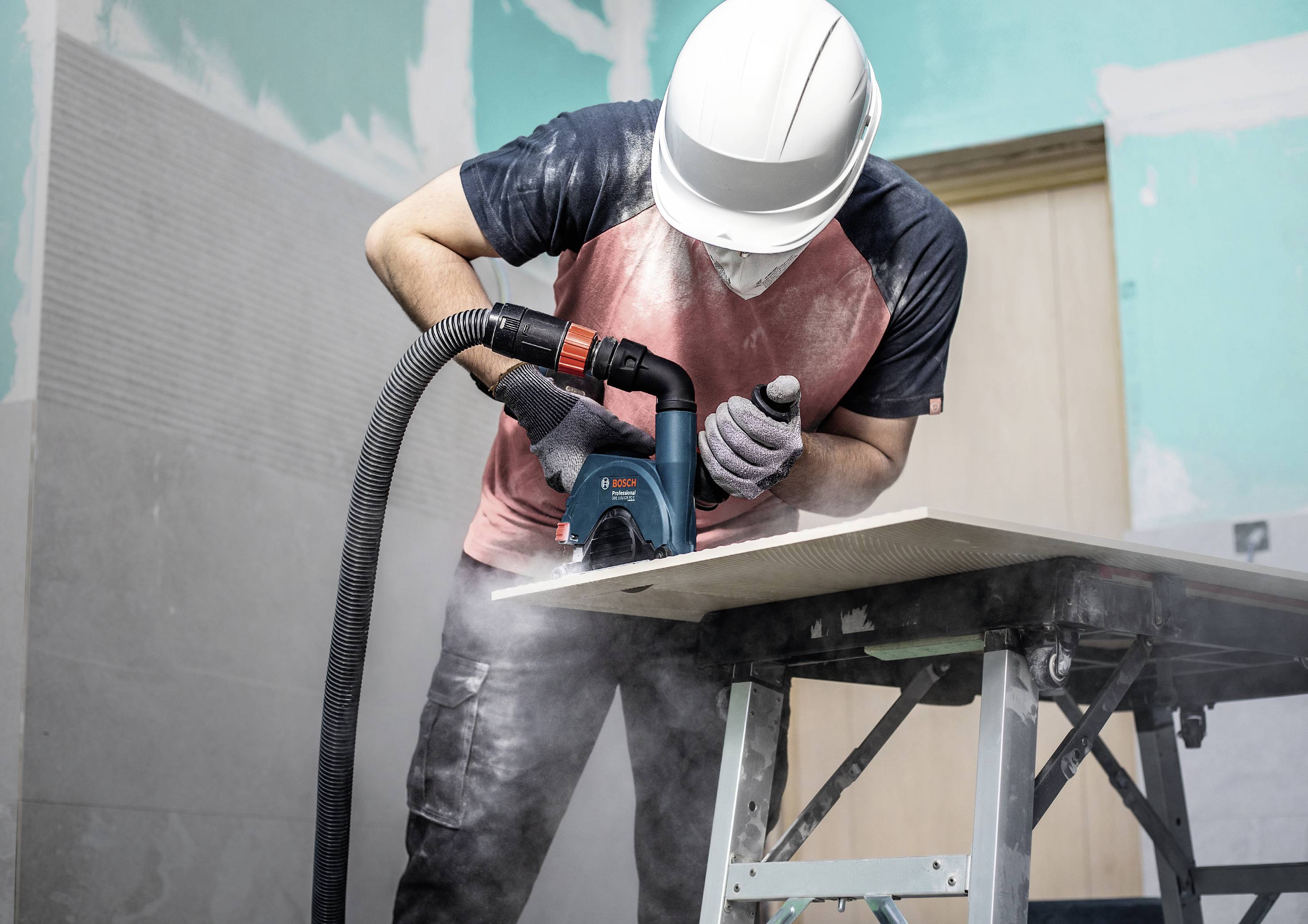 Person schneidet mit einer Maschine eine Holzplatte in einem Baustellenraum. Staub wird abgesaugt. Person trägt Schutzhelm und Handschuhe.