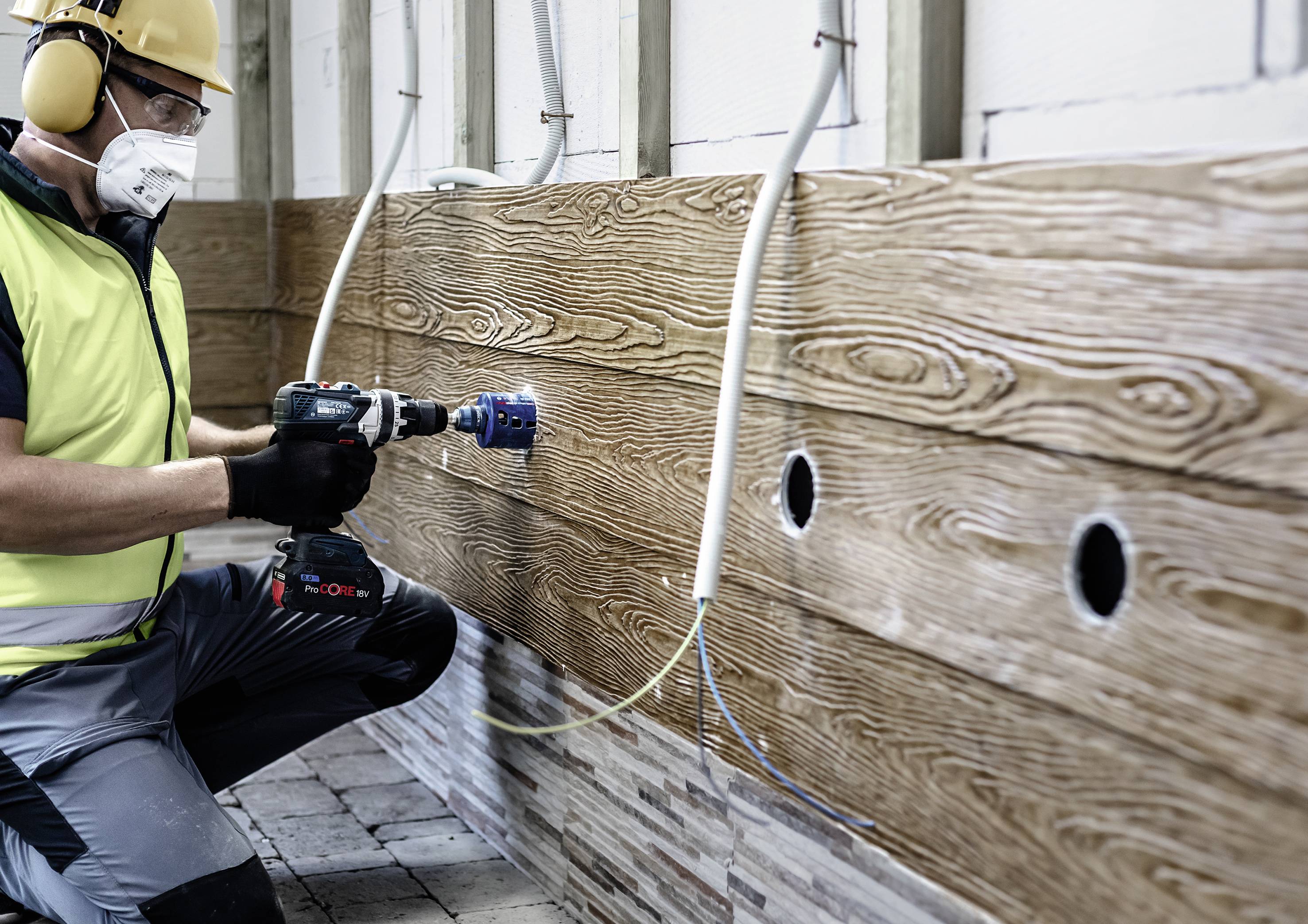 Ein Bauarbeiter in Schutzkleidung bohrt mit einer Maschine Löcher in eine Holzverkleidung. Kabel hängen lose aus der Wand.