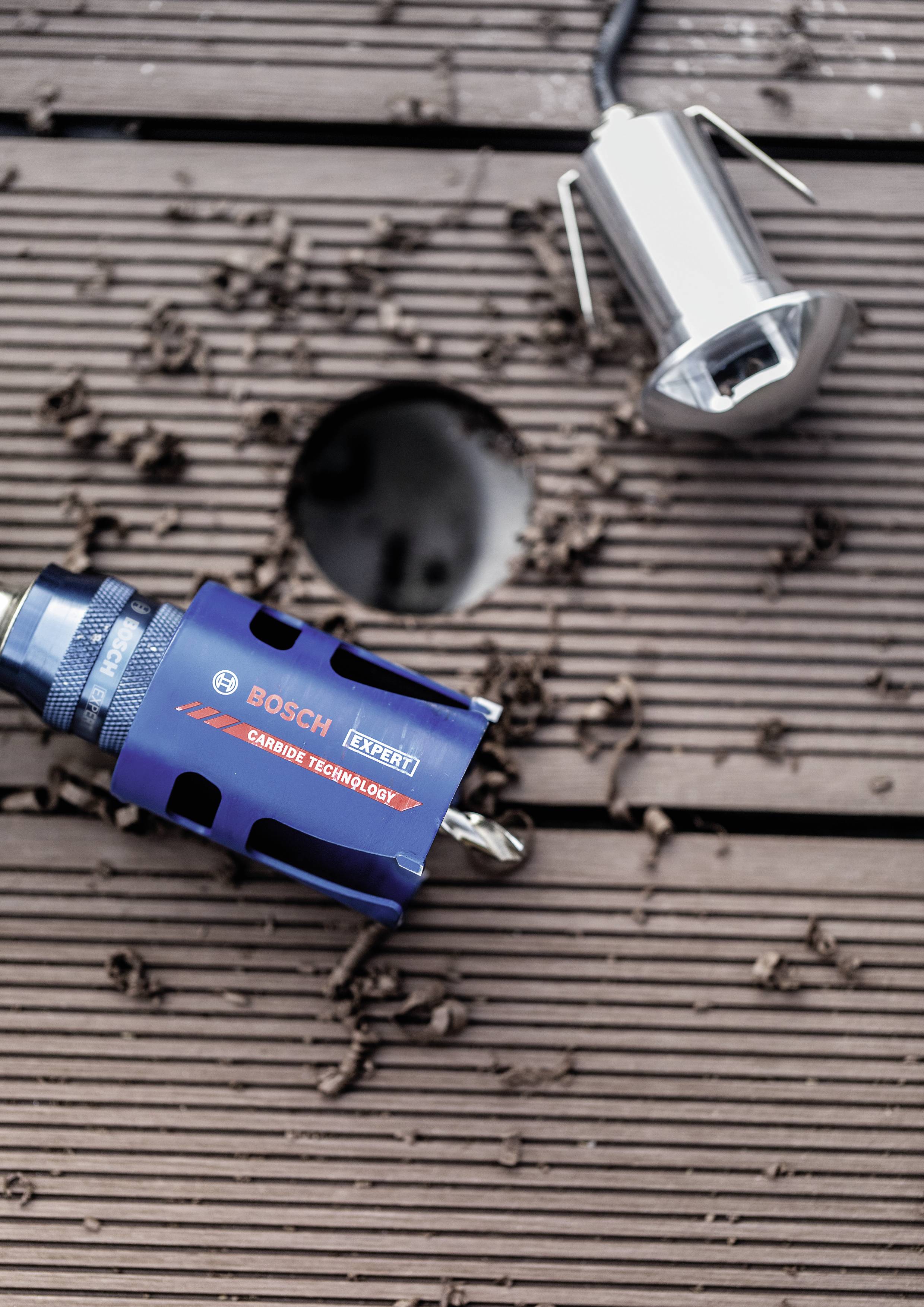 Ein blauer Bosch Lochsägeaufsatz liegt auf einem Holzboden neben einer Bohröffnung. Daneben liegt ein weiteres Metallstück.