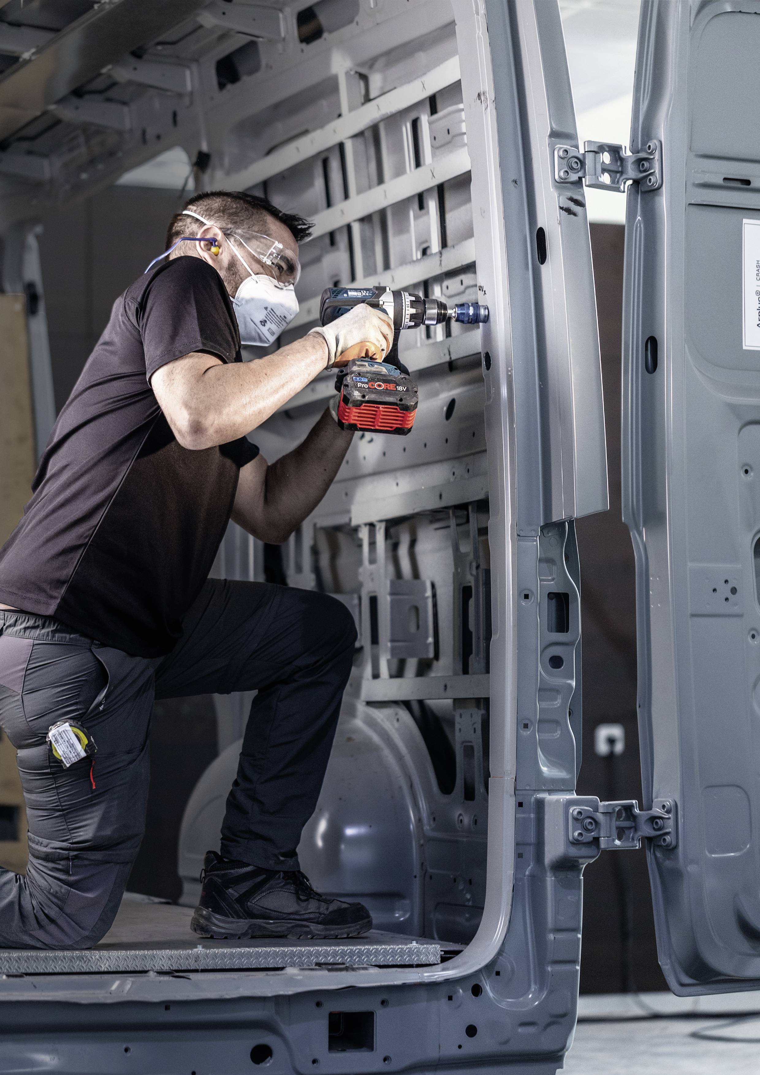 Ein Mann in Schutzmaske und Brille verwendet eine Bohrmaschine, um im Inneren eines Transporters zu arbeiten.