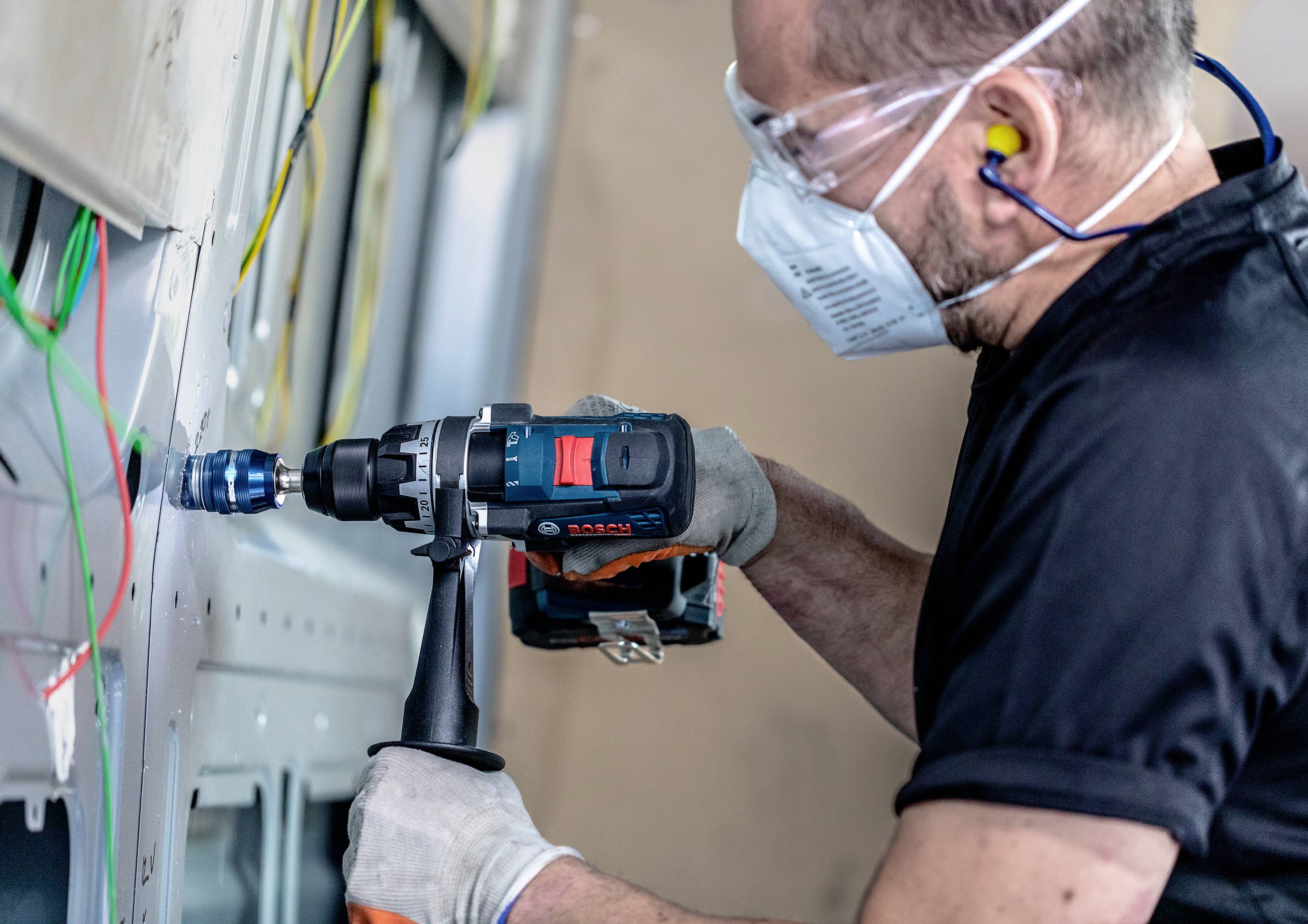 Ein Mann bohrt mit einer Bohrmaschine in eine Metallplatte. Er trägt Schutzbrille, Maske und Ohrstöpsel für den Arbeitsschutz.